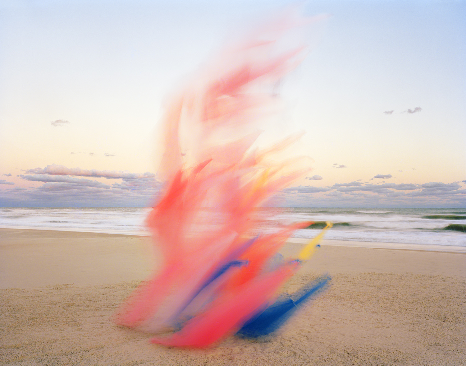 Blurred red and blue tulle fabric billows across a sandy beach at dusk, with gentle waves and a pastel sky visible in the soft-focus background.
