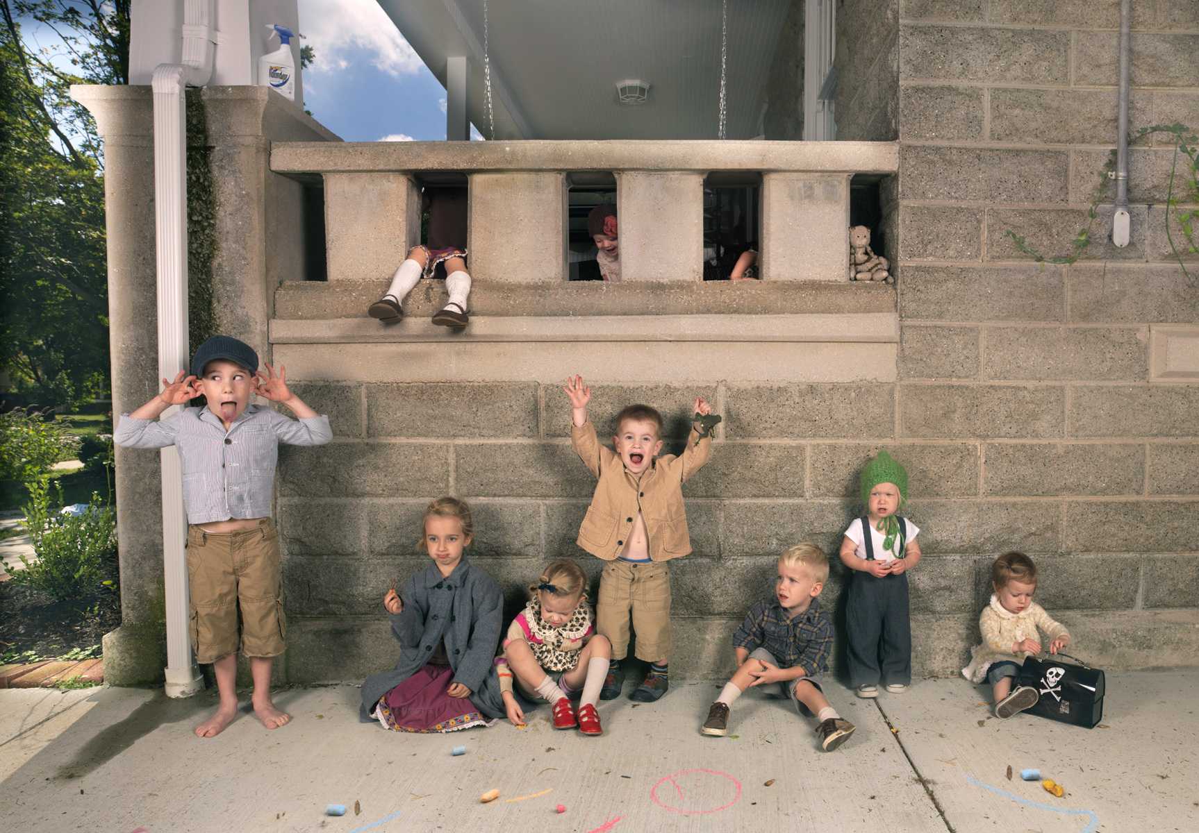 A group of children of varying ages stand and sit against a concrete brutalist building facade, arranged in a casual line. They wear vintage-style clothing in earth tones and pastels, with expressions of playful exuberance, while additional figures peer down from windows and openings above them.