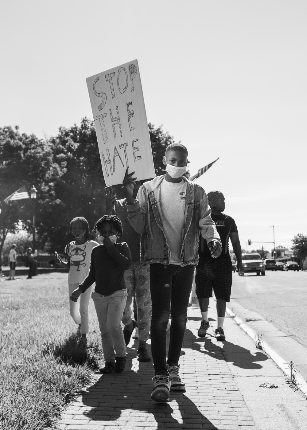 Black and white photograph of a group of people walking on a sidewalk, led by a young person wearing a face mask and holding a large sign reading "STOP THE HATE." Additional protesters, including children, walk alongside. Trees and parked cars are visible in the background.