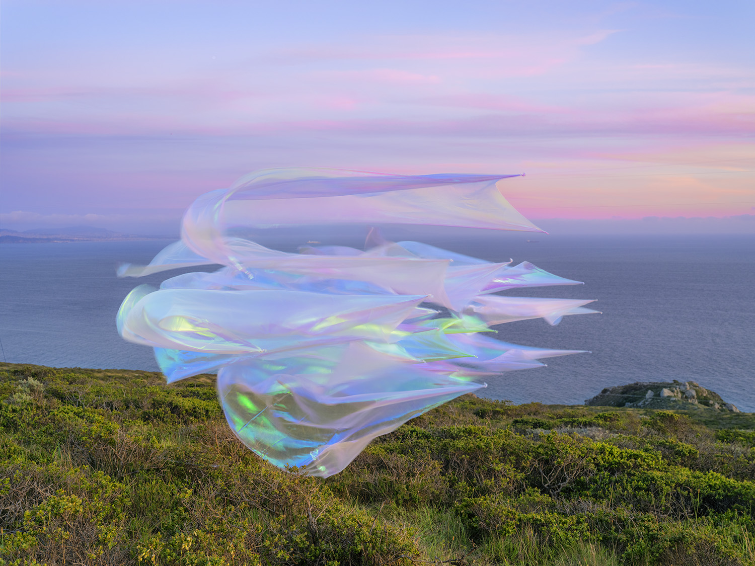 Iridescent, translucent fabric forms hover above a coastal hillside overlooking the ocean at dusk, their flowing surfaces catching pink, blue, and green light against the soft twilight sky.