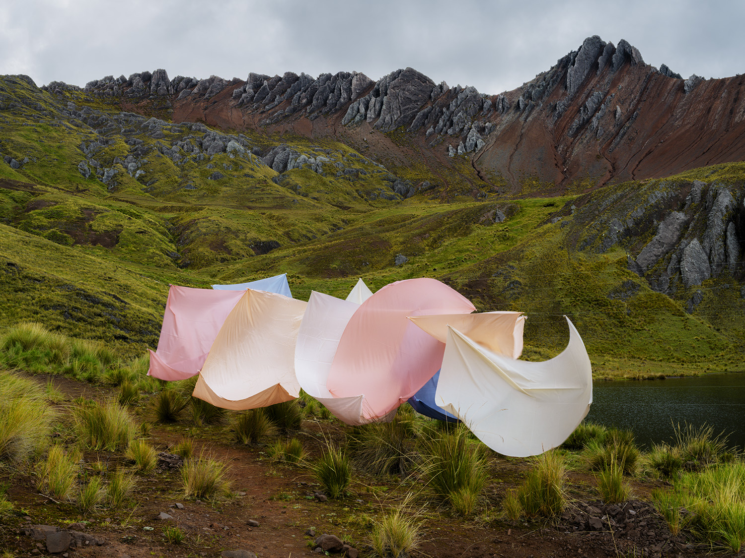 Billowing sheets of pink, cream, and white fabric suspended in a mountainous valley landscape, with moss-covered slopes, rocky outcroppings, and a small alpine lake visible beneath an overcast sky.