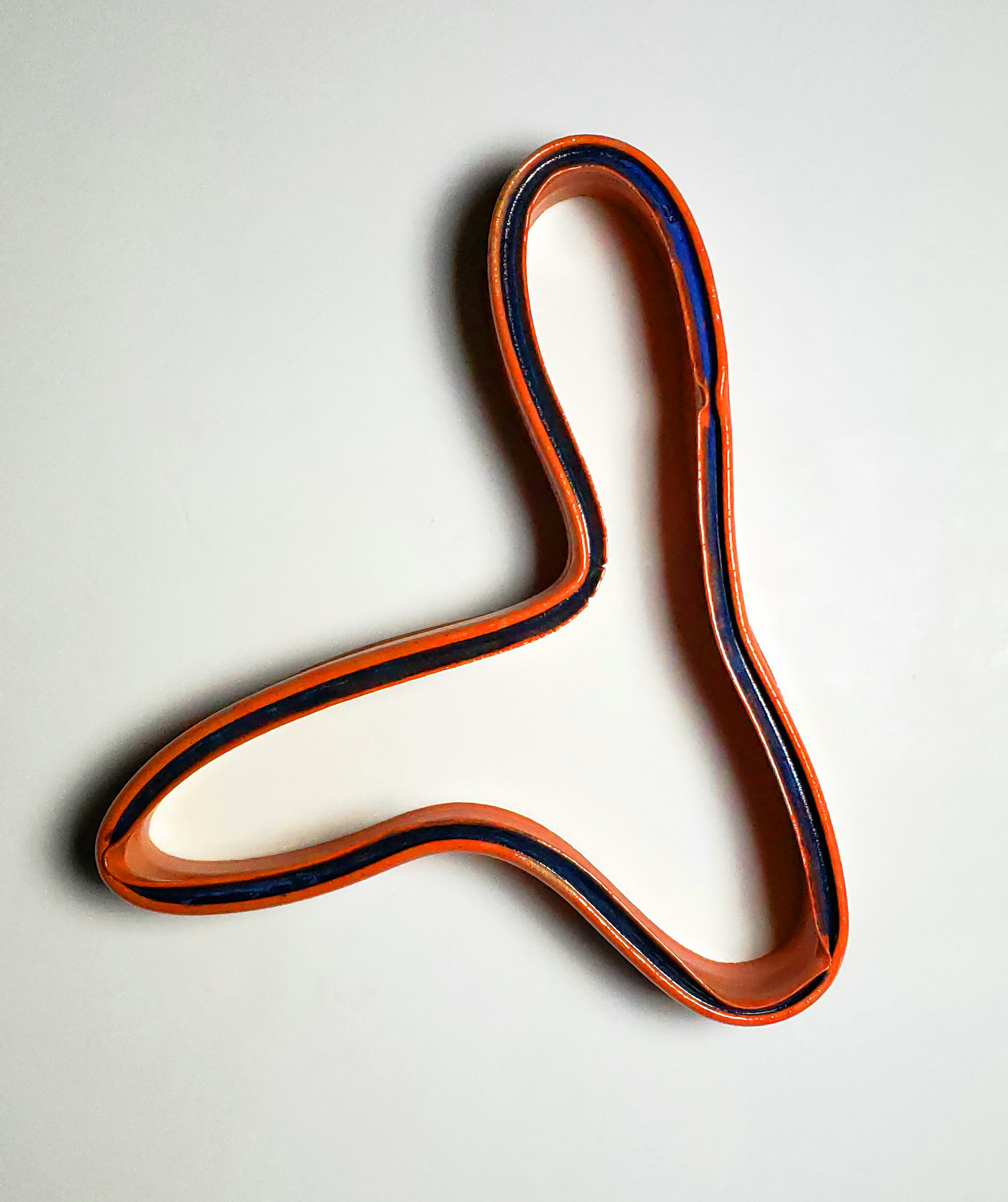 A continuous looping form in orange ceramic with a dark blue stripe running along its interior edge, photographed against a white background. The sculptural piece creates two interconnected oval shapes with graceful, organic curves.