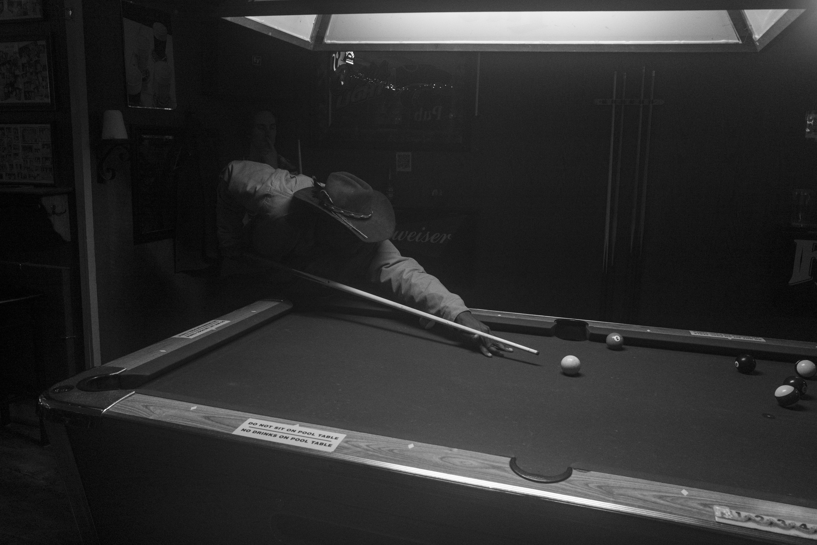 Black and white photograph of a person leaning over a pool table mid-shot, cue in hand, with billiard balls scattered across the felt surface. The composition captures the interior of what appears to be a dimly lit recreation space, with overhead lighting and signage visible in the background.