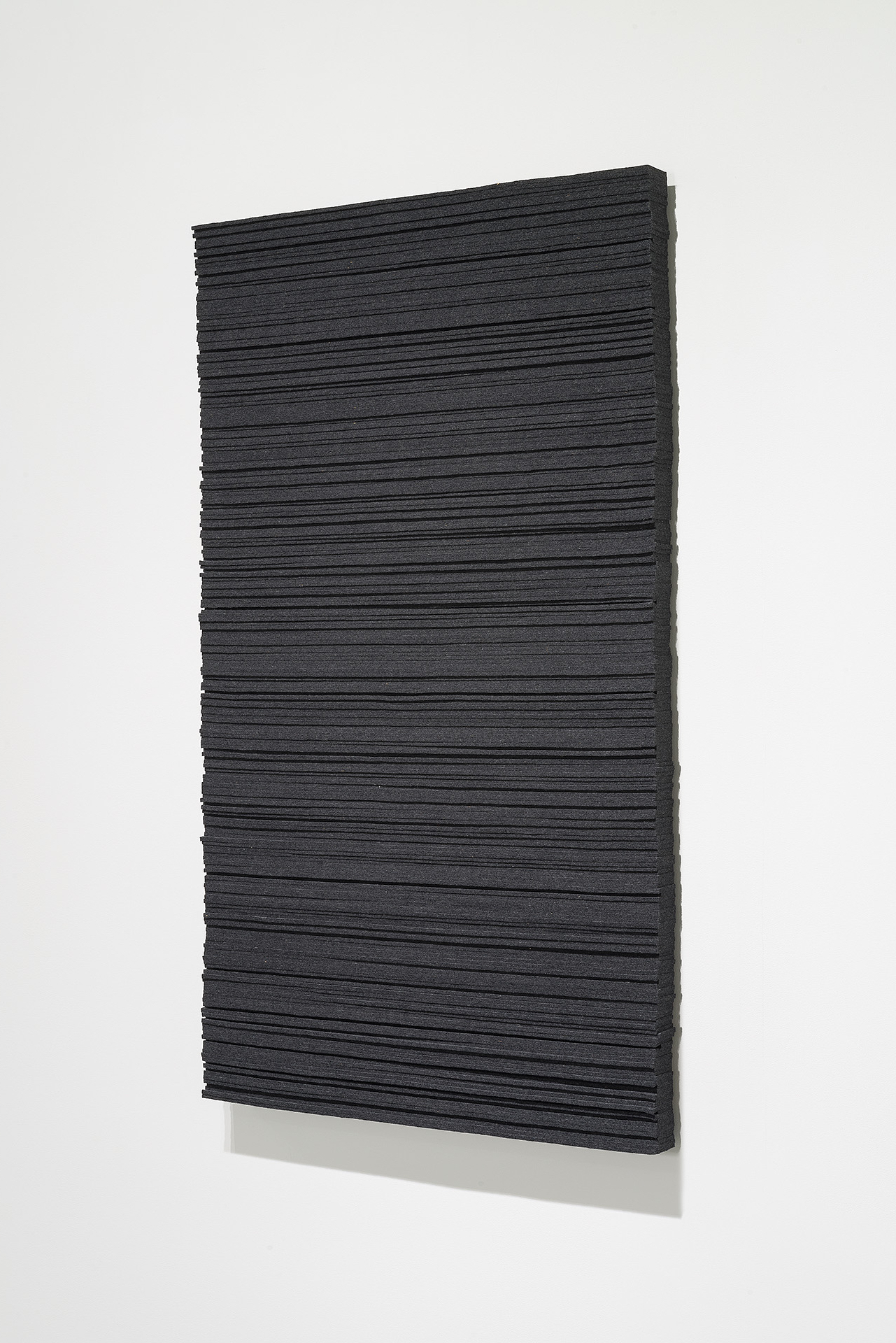 A rectangular panel of dark charcoal wool felt with horizontal linear ridges across its surface, mounted at a slight angle against a light gray background with a pale base visible beneath.