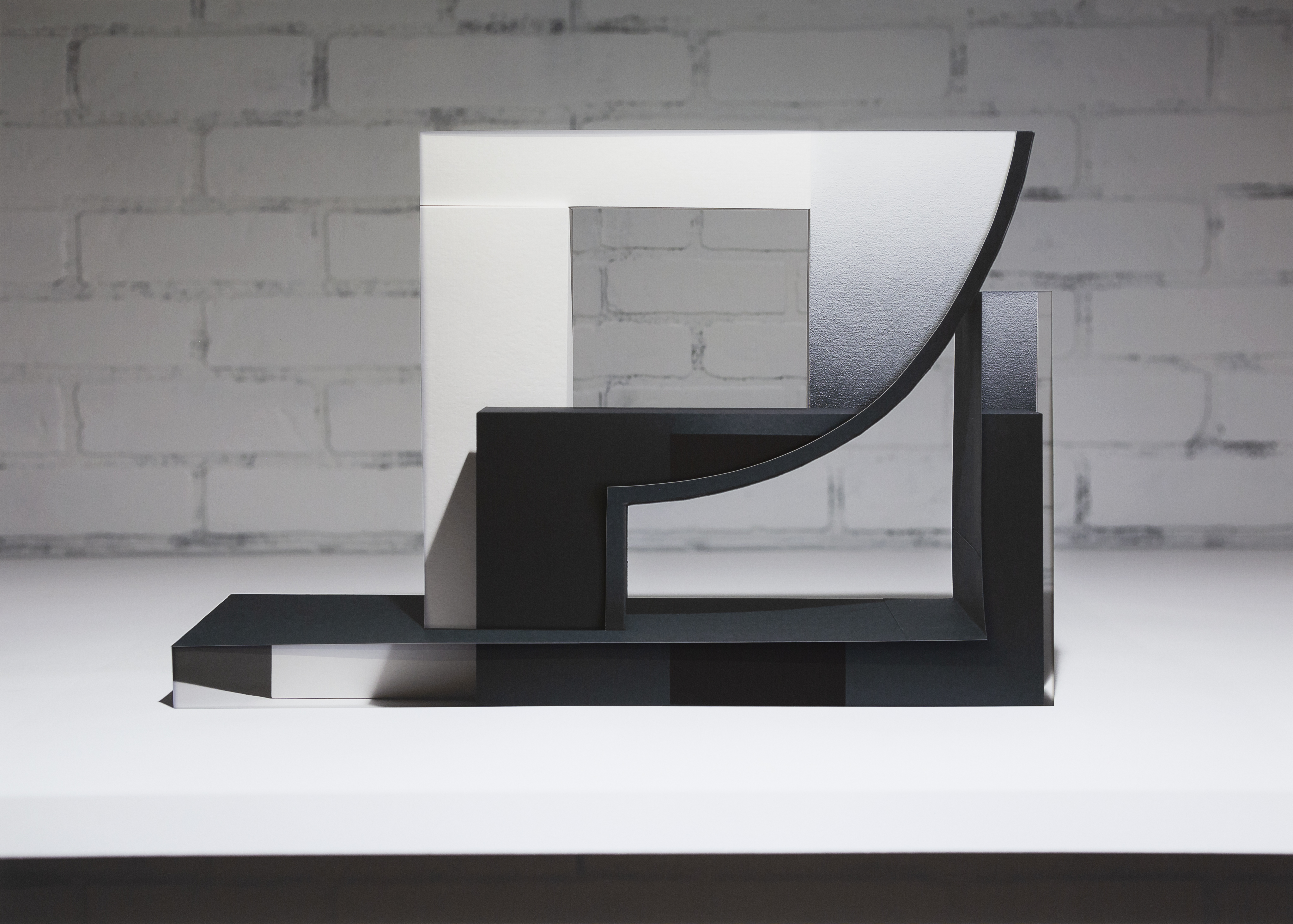 Abstract black sculptural form composed of geometric planes and curved elements, displayed against a white gallery wall with visible brick texture. The composition features layered, translucent paper elements in white and gray tones creating depth and dimensionality through collage and decollage techniques.