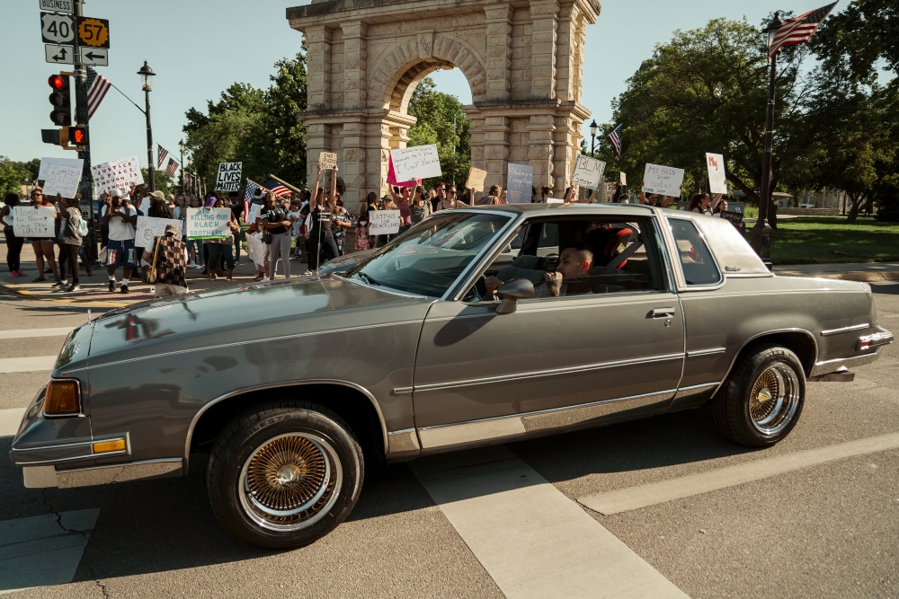 A gray sedan with gold-trimmed wheels drives through a city street lined with protesters holding signs. Behind the vehicle, a large stone arch monument is visible, with American flags and crowds gathered in the background under clear sky.