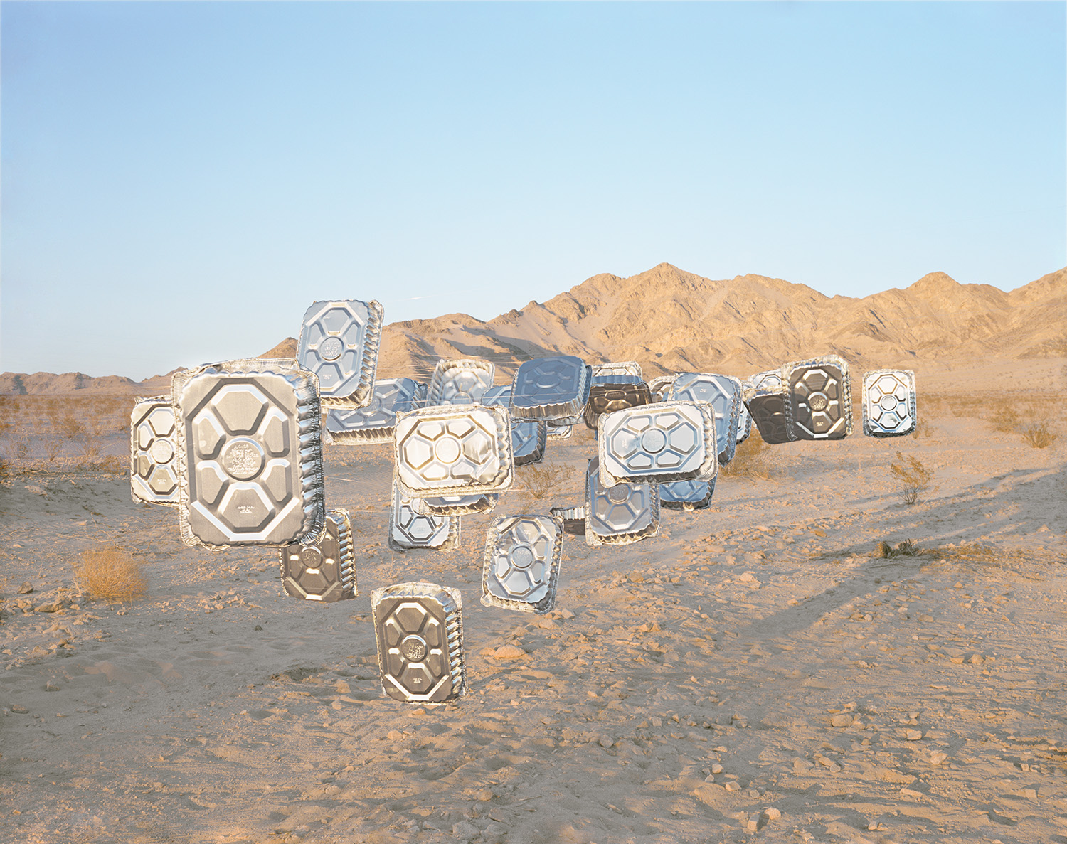 Sculptural installation of metallic geometric forms with hexagonal and circular patterns arranged in a line across a desert landscape, with sandy hills visible in the background under a clear sky.