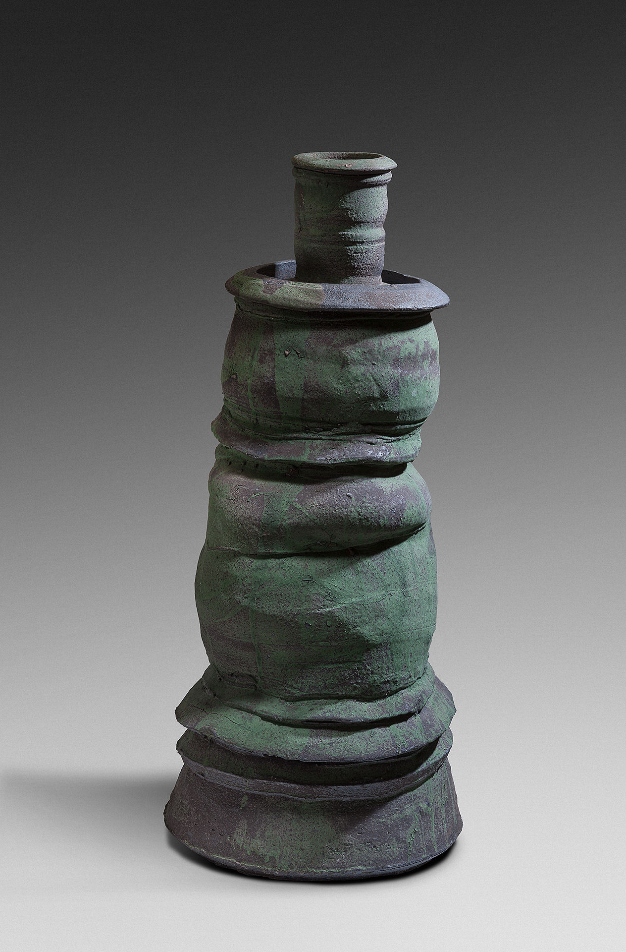 A stacked ceramic vessel in dark greenish-black stoneware with a weathered patina. The sculptural form is composed of bulbous, nested sections that taper toward a cylindrical neck topped with a flat cap, creating an unstable, gravity-defying composition that appears to slump and shift under its own weight.