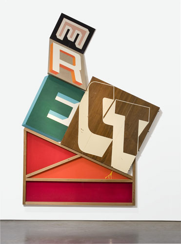 A tilted wooden house-shaped construction with stacked, colorful letters spelling "ERECT" in various typefaces and hues including orange, green, tan, brown, and red. The structure leans dramatically to one side, with geometric shapes and bold typography creating a playful, off-balance composition against a neutral background.