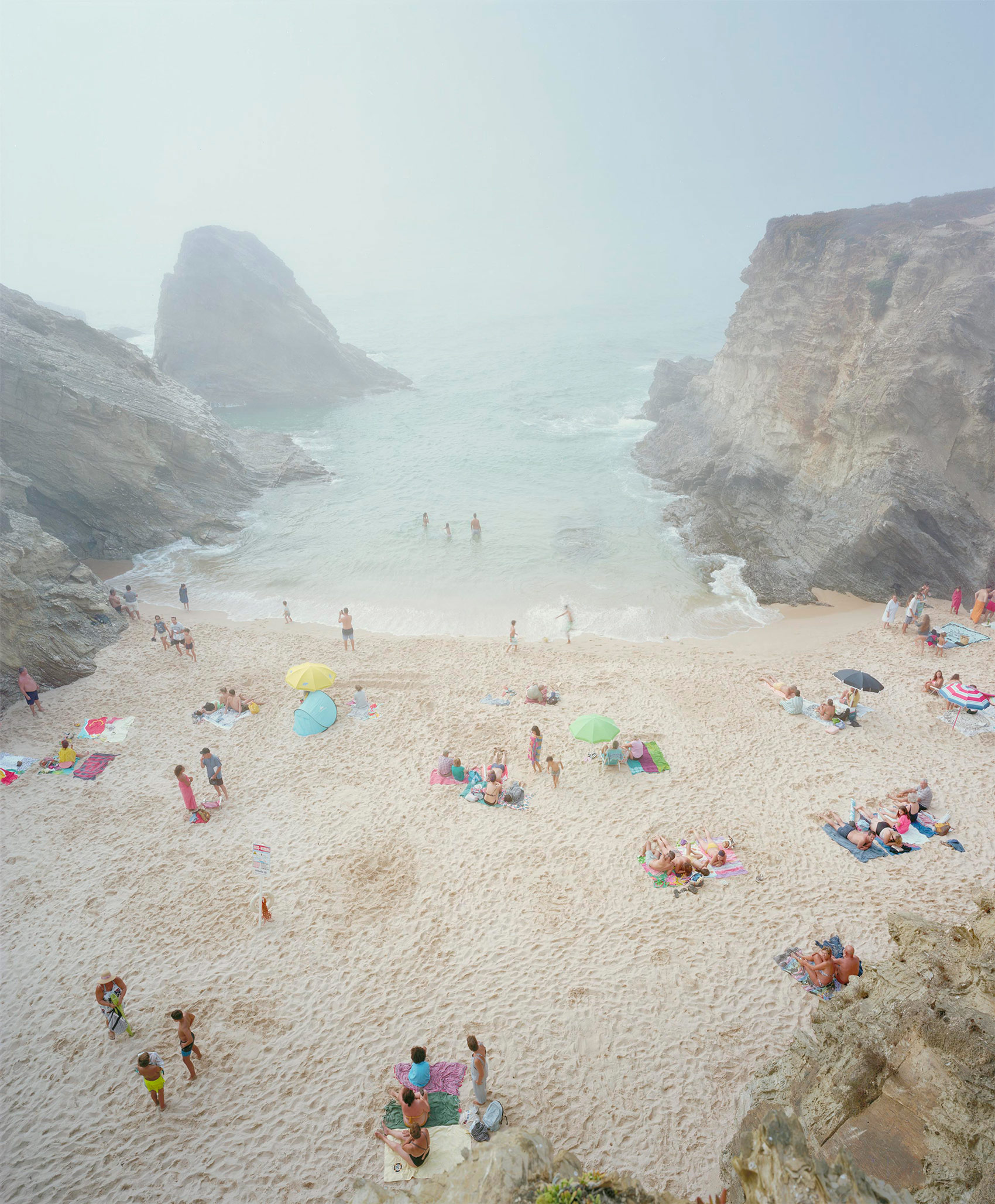 Aerial view of a secluded beach cove nestled between dramatic cliffsides, with scattered beachgoers, colorful umbrellas, and towels dotting the pale sand. Turquoise shallow waters and a large rocky sea stack visible in the misty distance.