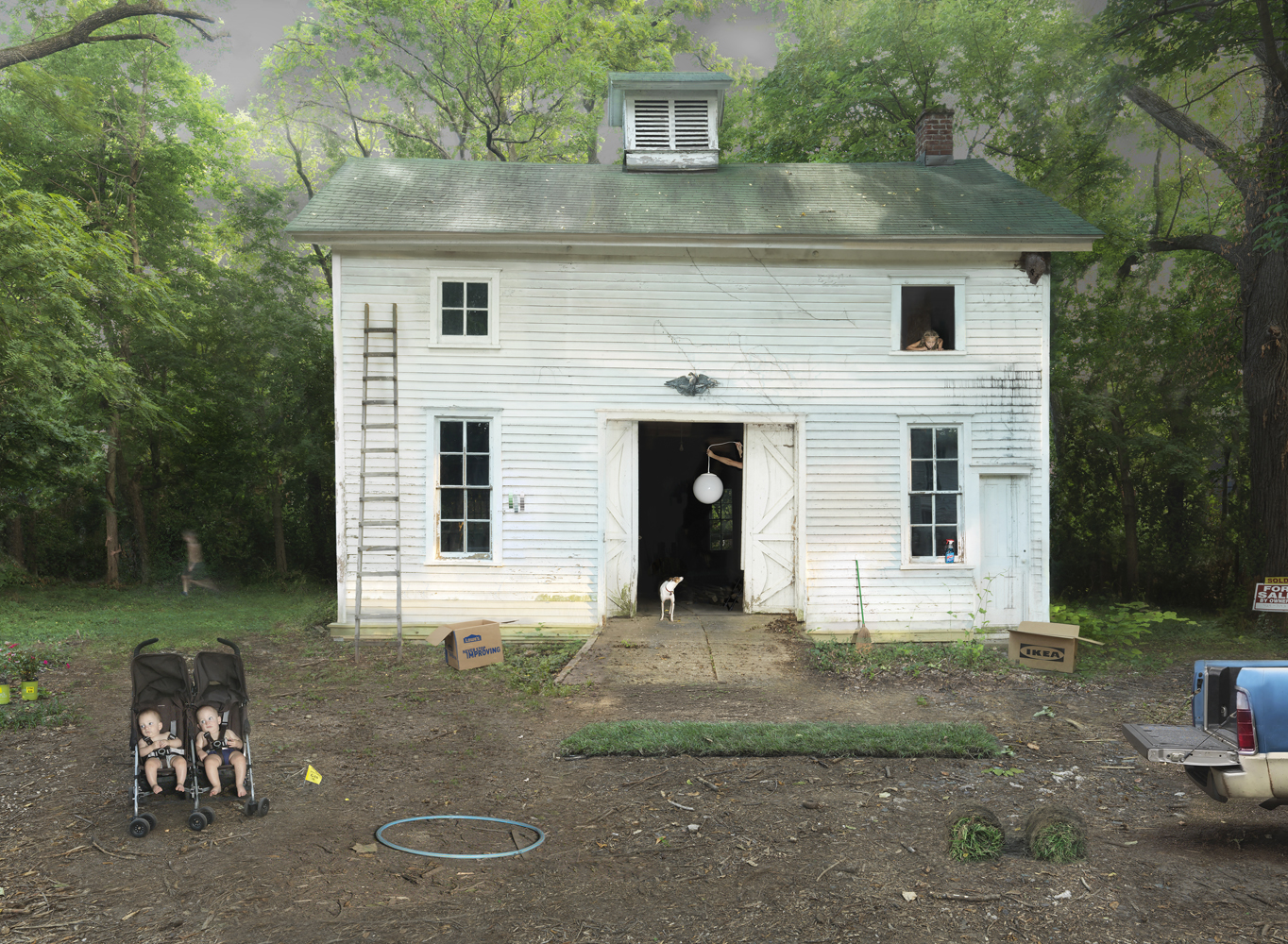 A white wooden house surrounded by dense green trees sits in a dirt yard scattered with children's toys, including a double stroller on the left, a hula hoop, and cardboard boxes. Figures appear in several windows and doorways of the modest two-story structure, while a small dog stands near the open front entrance. The scene is composed as a centered, frontal view with a documentary-like quality.