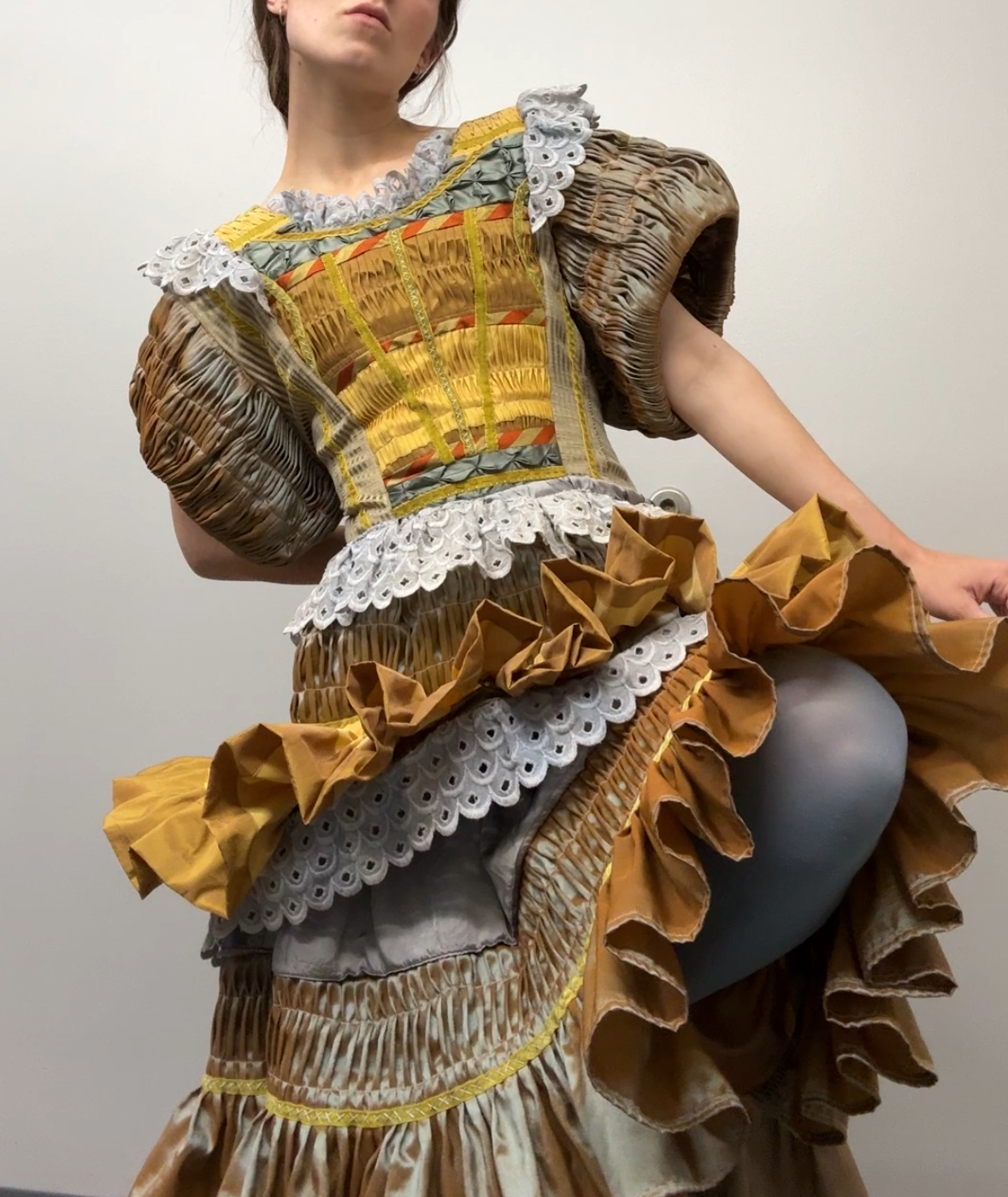 A figure wearing an elaborate dress constructed from pleated and embroidered taffeta and cotton in gold, yellow, and cream tones. The garment features voluminous puffed sleeves, intricate white lace trim, layered ruffles, and detailed pleating throughout, creating a sculptural silhouette against a neutral background.