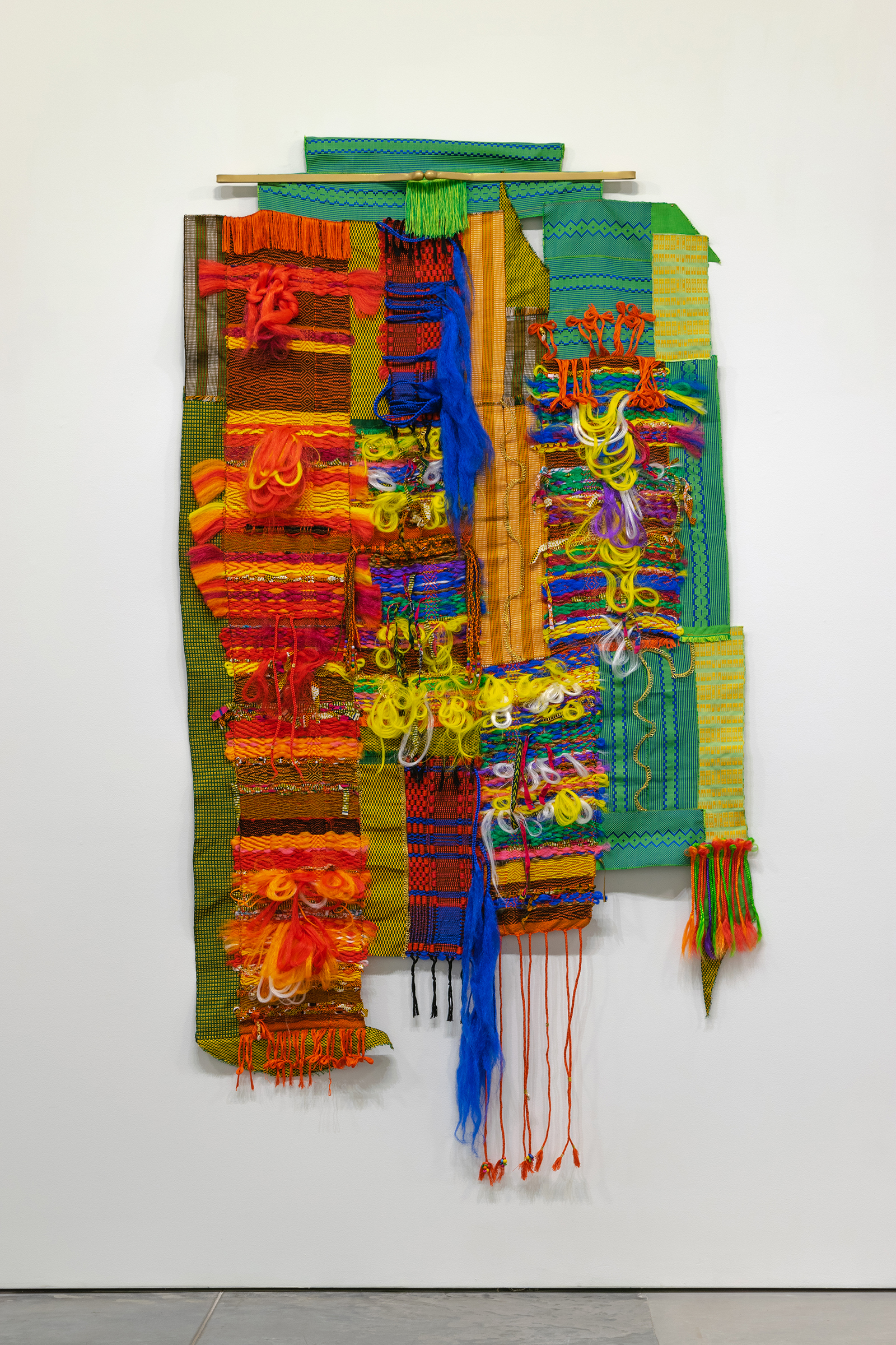 Vibrant textile wall hanging composed of layered fabric strips in shades of red, orange, yellow, green, and blue, incorporating woven patterns, fringe elements, and dangling decorative fibers. The asymmetrical composition features contrasting textures and materials arranged vertically against a white gallery wall.