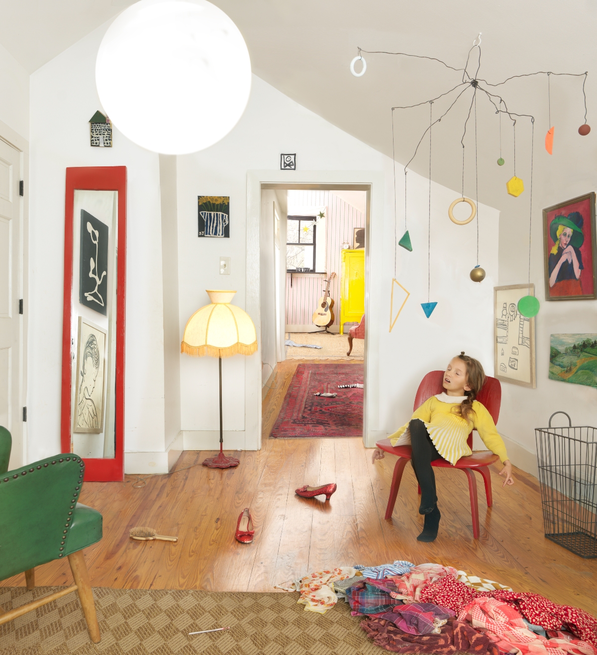 A young girl in a yellow sweater sits in a red chair gazing upward in a bright, eclectic interior space. The room features white walls, natural wood flooring, colorful artworks and objects including a large illuminated ceiling fixture, a hanging mobile with geometric shapes and ornaments, a yellow-fringed floor lamp, and scattered clothing on the floor, creating a whimsical, lived-in domestic scene filled with vibrant reds, greens, yellows, and blues.
