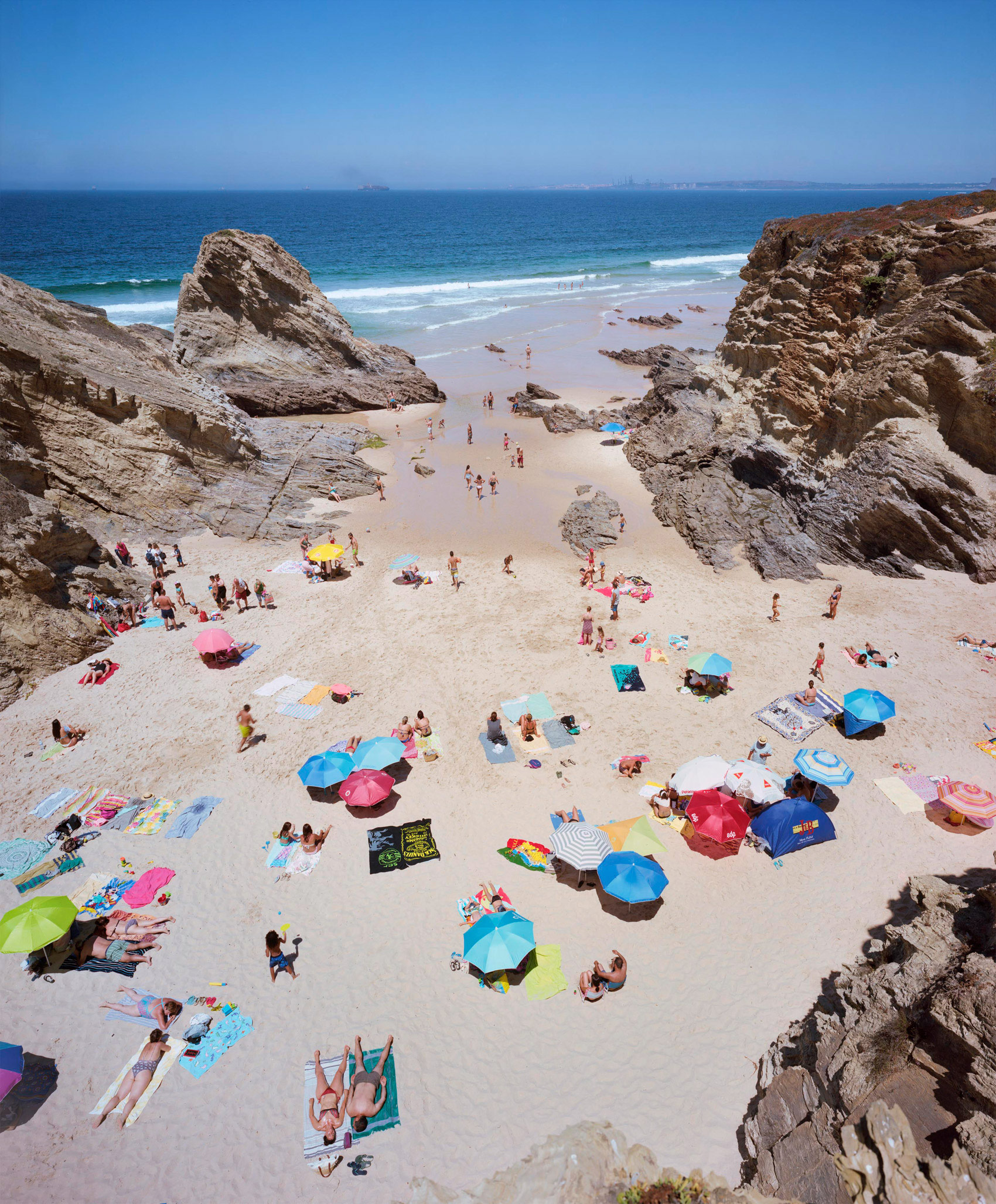 Aerial view of a crowded beach nestled between dramatic rocky outcrops, with sunbathers scattered across pale sand beneath colorful umbrellas and beach towels in shades of blue, pink, red, and yellow. Clear turquoise waters and white-capped waves frame the composition under a cloudless sky.