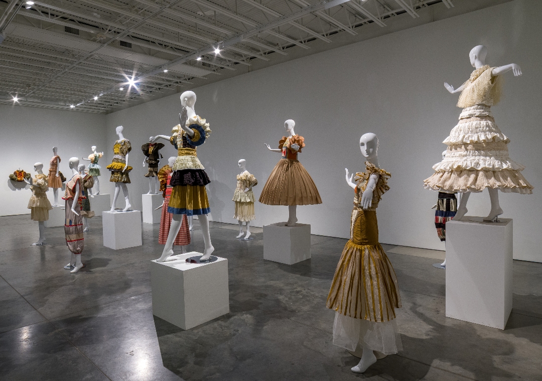 Gallery installation view featuring approximately fifteen mannequins dressed in elaborately constructed garments displayed on white pedestals. The pieces showcase hand-pleated skirts, embroidered details, and layered fabrics in warm tones of gold, bronze, cream, and burgundy, with sculptural silhouettes ranging from voluminous tiered skirts to structured bodices, set against white walls under gallery lighting.