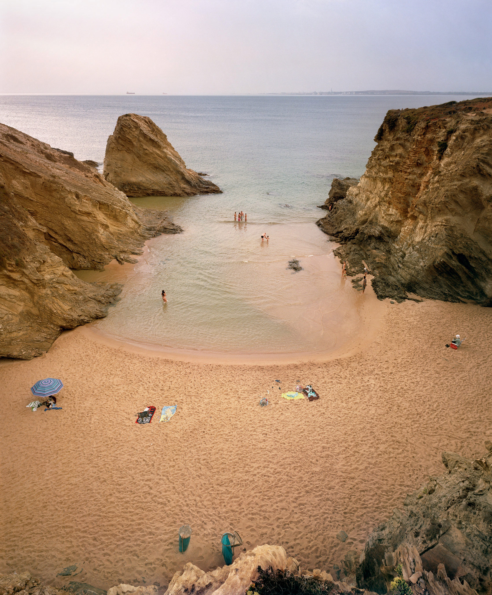 A secluded cove with sandy beach framed by dramatic golden-brown rock formations on either side. Several beachgoers with umbrellas and belongings are scattered across the sand, while others wade in the pale turquoise shallow water. The distant horizon shows calm sea and a hazy shoreline, creating a serene coastal landscape captured in warm, naturalistic tones.