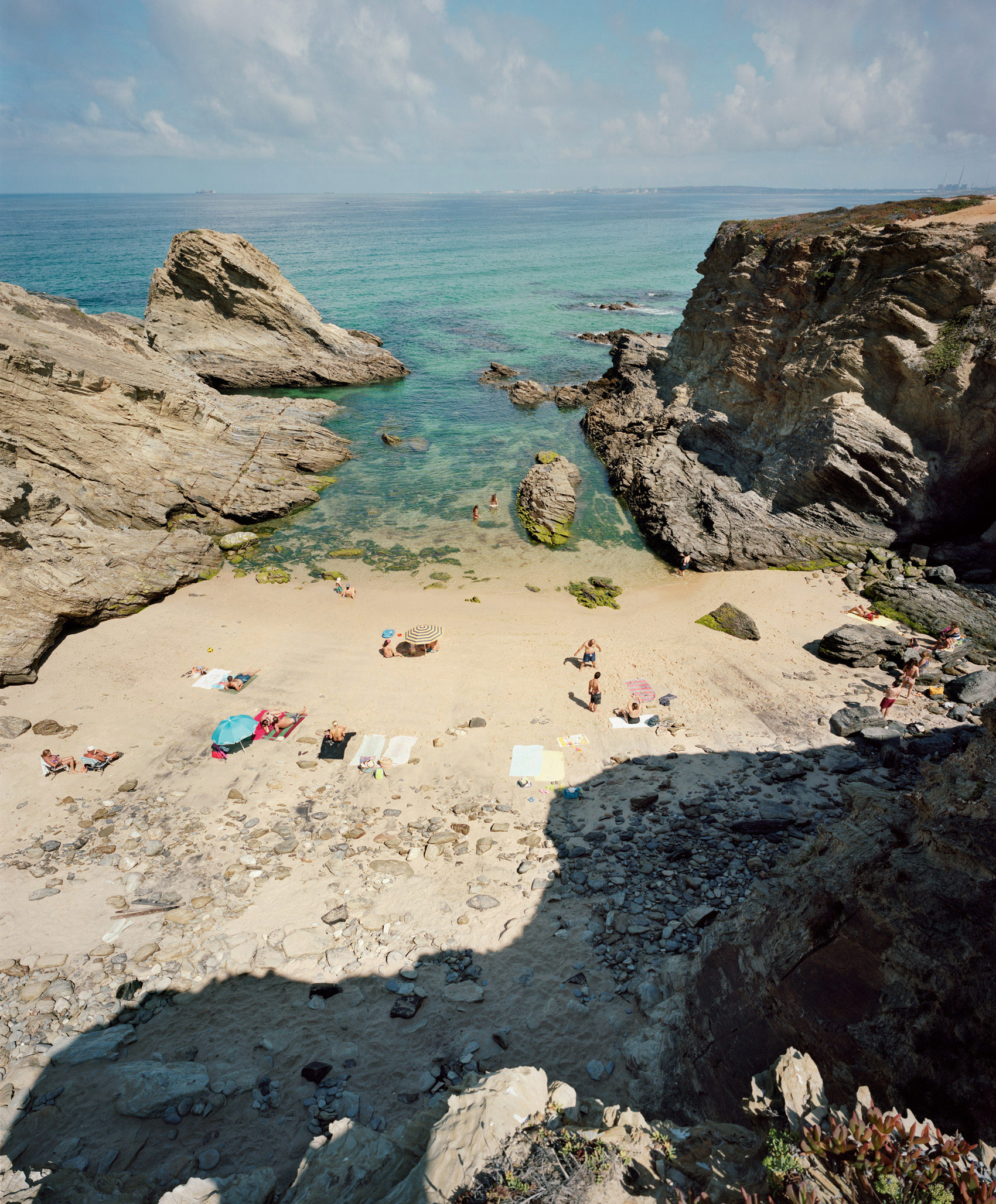 Aerial view of a secluded sandy beach nestled between dramatic rocky outcrops and turquoise water, with scattered beachgoers and colorful umbrellas dotting the pale sand below.