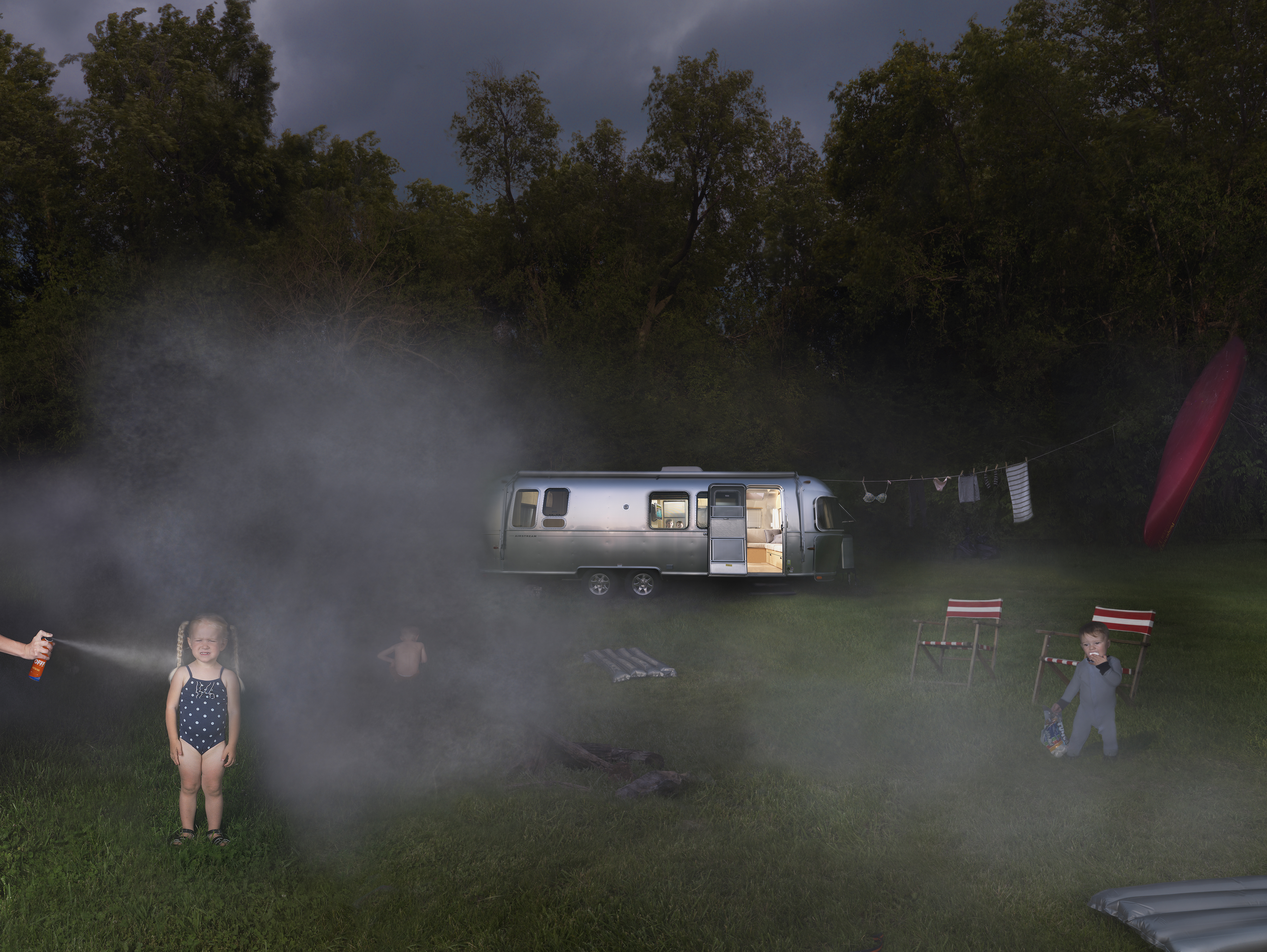 A nighttime campsite scene shows children playing in a misty field before an illuminated silver Airstream trailer, with folding chairs and trees visible through the hazy atmosphere.
