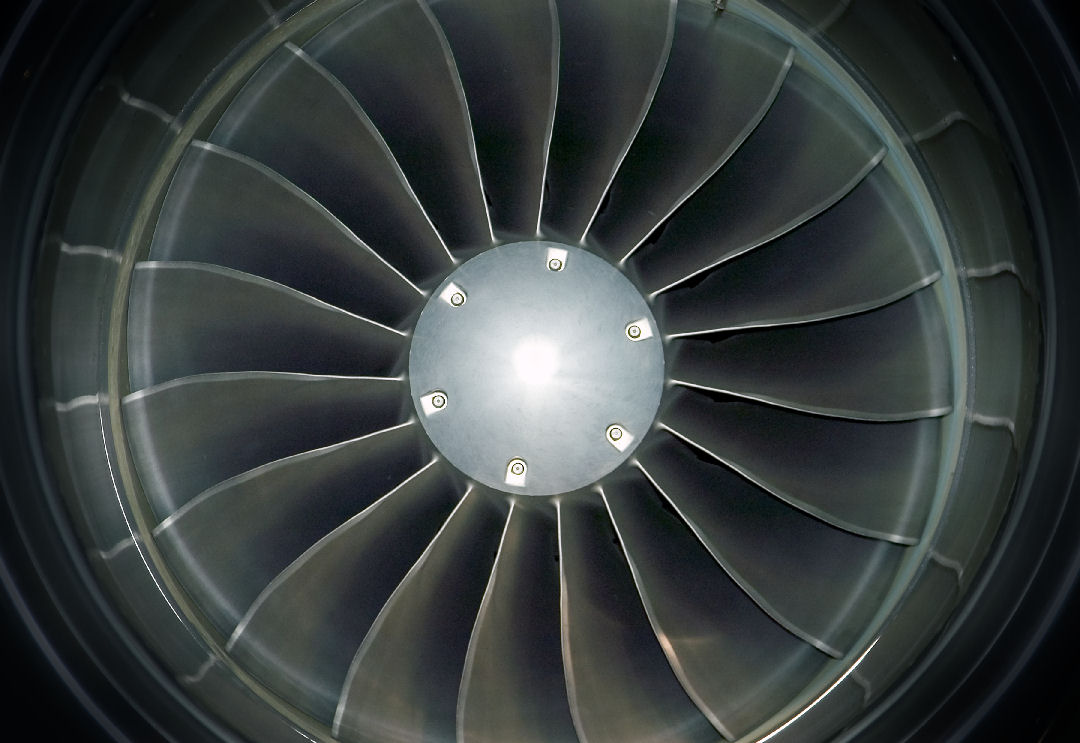 Frontal view of a jet engine turbine with multiple dark metallic blades radiating from a central silver hub, mounted within a circular housing. The geometric composition emphasizes symmetry and industrial precision through the fan's repetitive blade structure.
