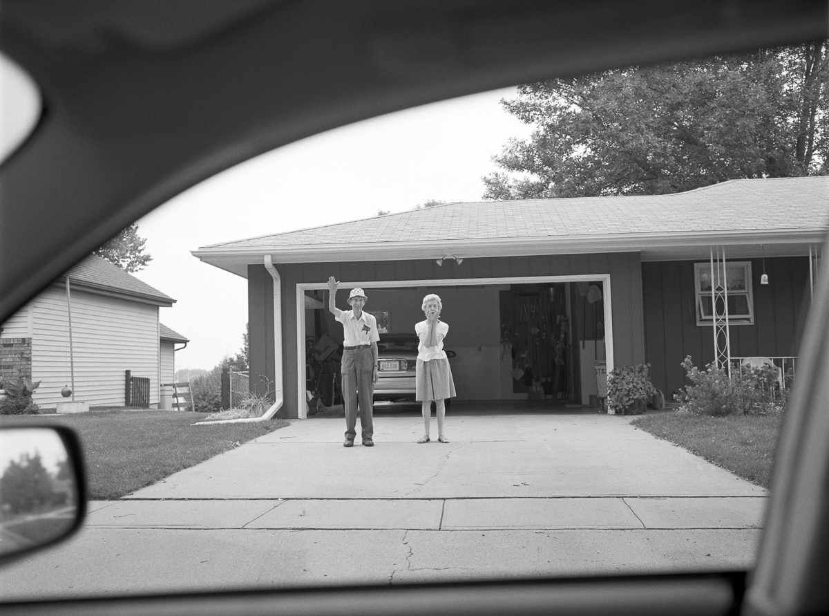 Black and white photograph taken from inside a car, framed by the windshield and side mirror, showing a man and woman standing in a residential driveway before a single-story house with an open garage. The couple, dressed in mid-century clothing, wave toward the camera as a vintage automobile sits in the garage behind them.