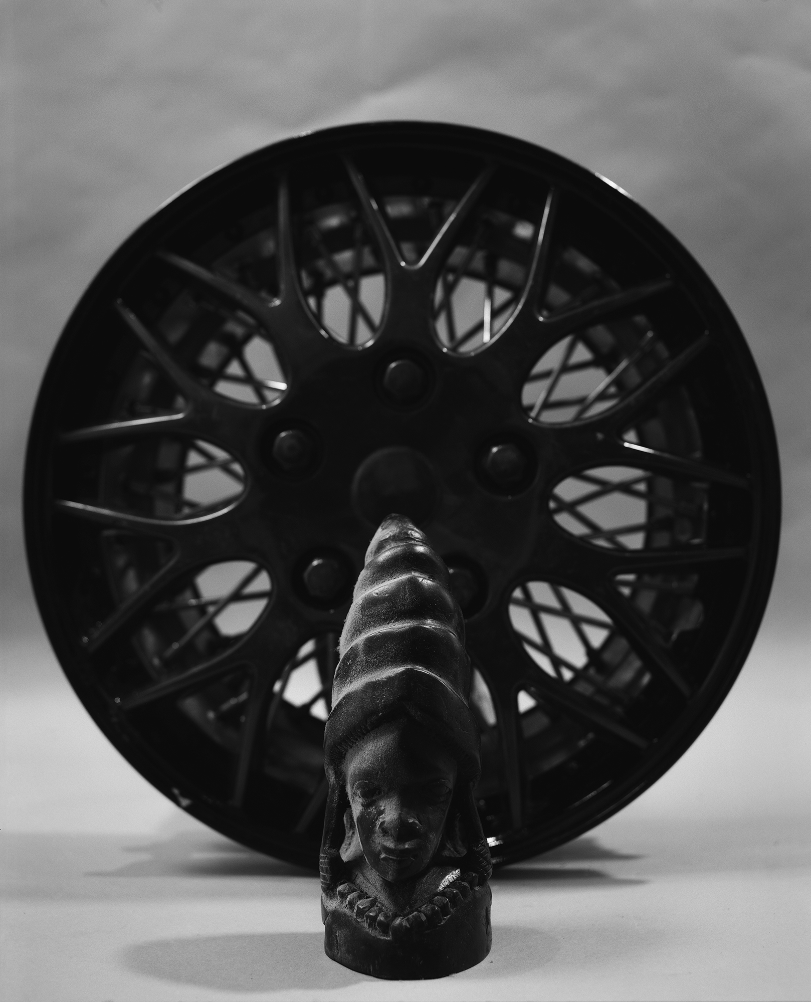 A black and white still life composition featuring a carved wooden bust with an ornate collar in the foreground, positioned before a large ornamental wheel with intricate spoke patterns. The geometric forms create strong contrasts of light and shadow against a neutral background.