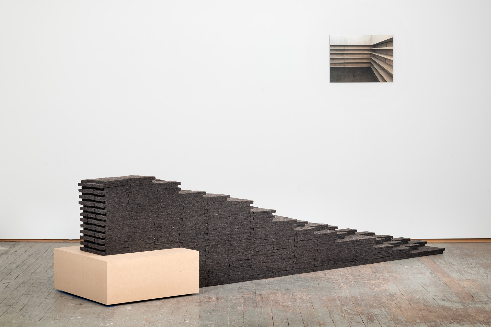 A sculptural installation featuring stacked dark charcoal wool felt panels arranged in a descending stepped formation across a wooden gallery floor, rising from a cream-colored rectangular base. A framed photograph of similar stepped architectural forms hangs on the white wall above.