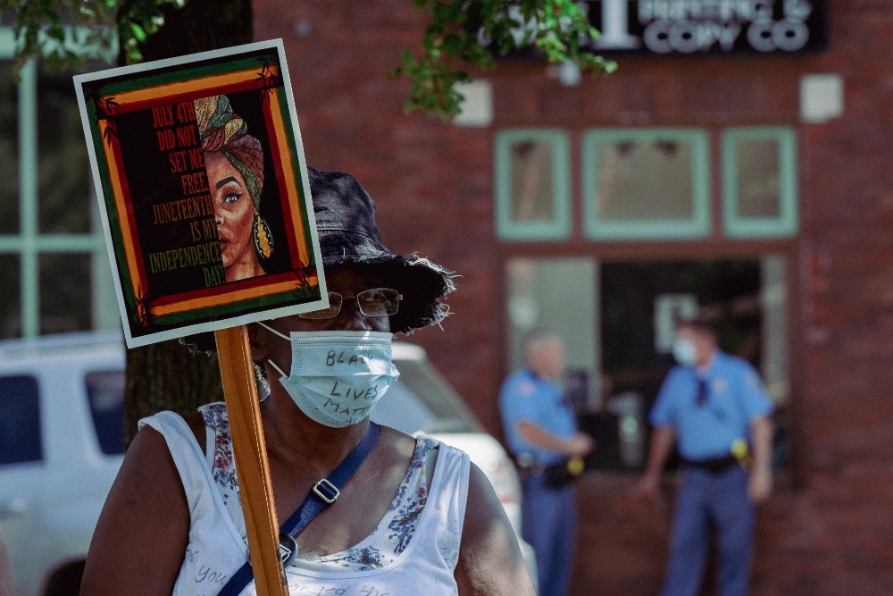 A protester wearing a face mask holds a wooden sign featuring a portrait of a woman's face in warm orange and red tones with text overlaid, standing before a brick building with uniformed officers visible in the background.