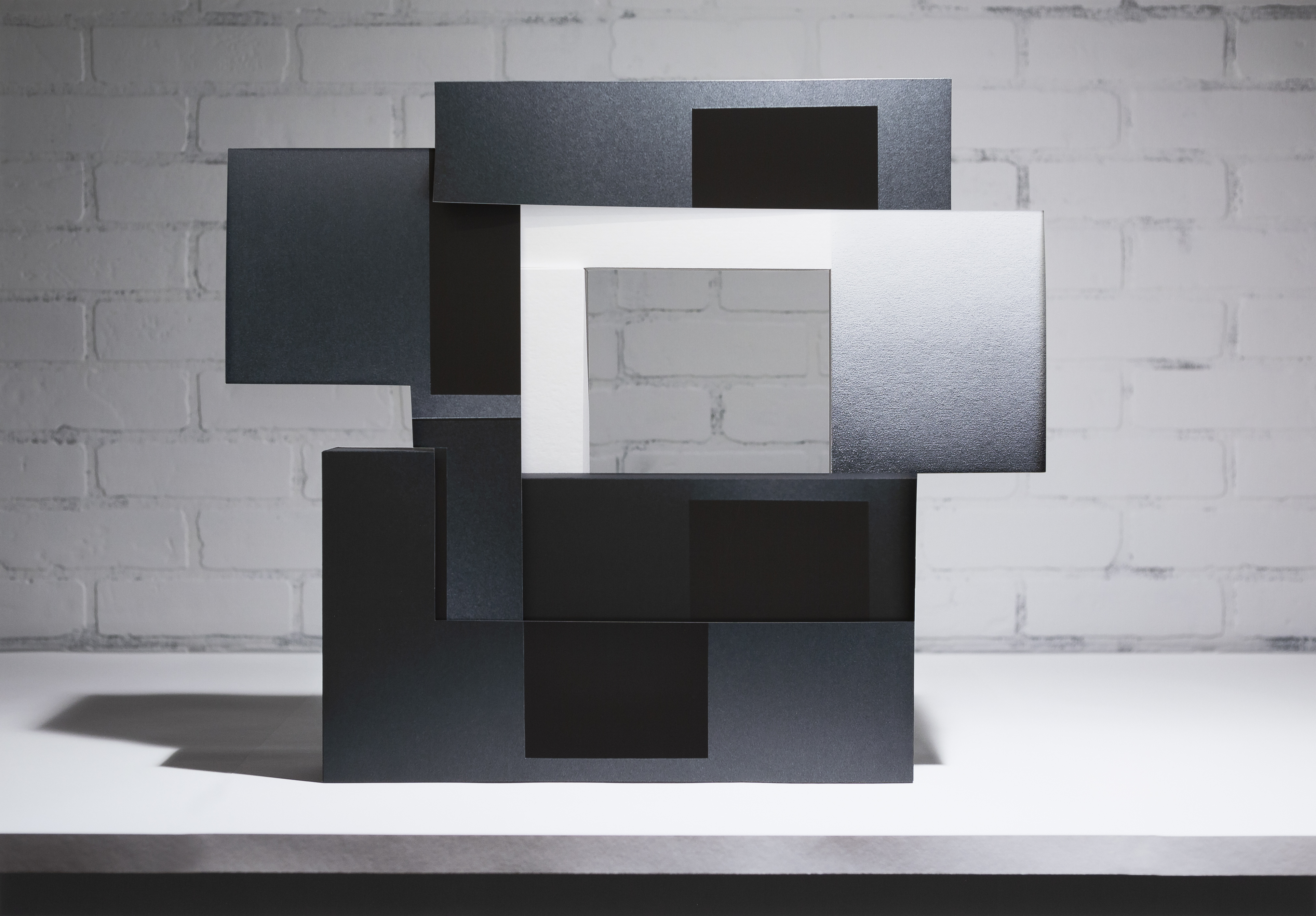 Layered rectangular panels in black, dark gray, and white arranged in an overlapping, three-dimensional composition against a white brick wall. The stacked forms create depth through varying planes and tones, with some surfaces appearing textured or printed, casting subtle shadows on the white surface beneath.