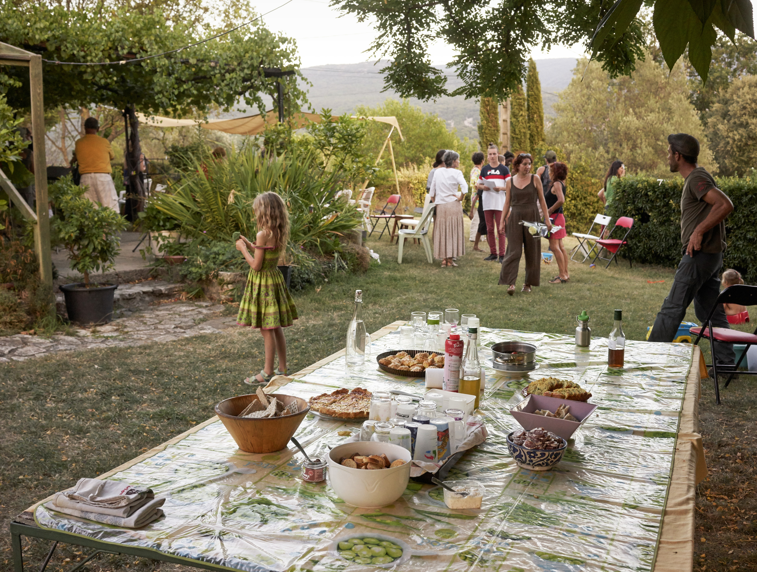 A garden gathering in a sunlit Tuscan-style landscape, with a long outdoor table laden with food and bottles in the foreground, and multiple guests mingling across a terraced lawn scattered with chairs and potted plants, mountains visible in the hazy distance.