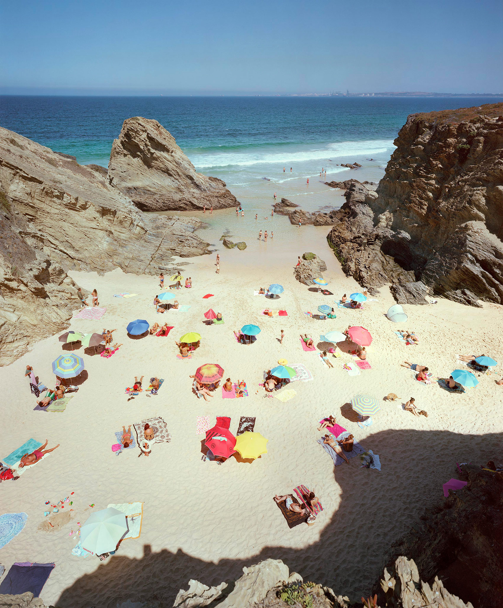 Aerial view of a crowded beach nestled between dramatic rocky outcrops, with numerous colorful umbrellas and towels dotting the pale sand. Turquoise and blue waters with gentle waves extend to the horizon under clear sky.