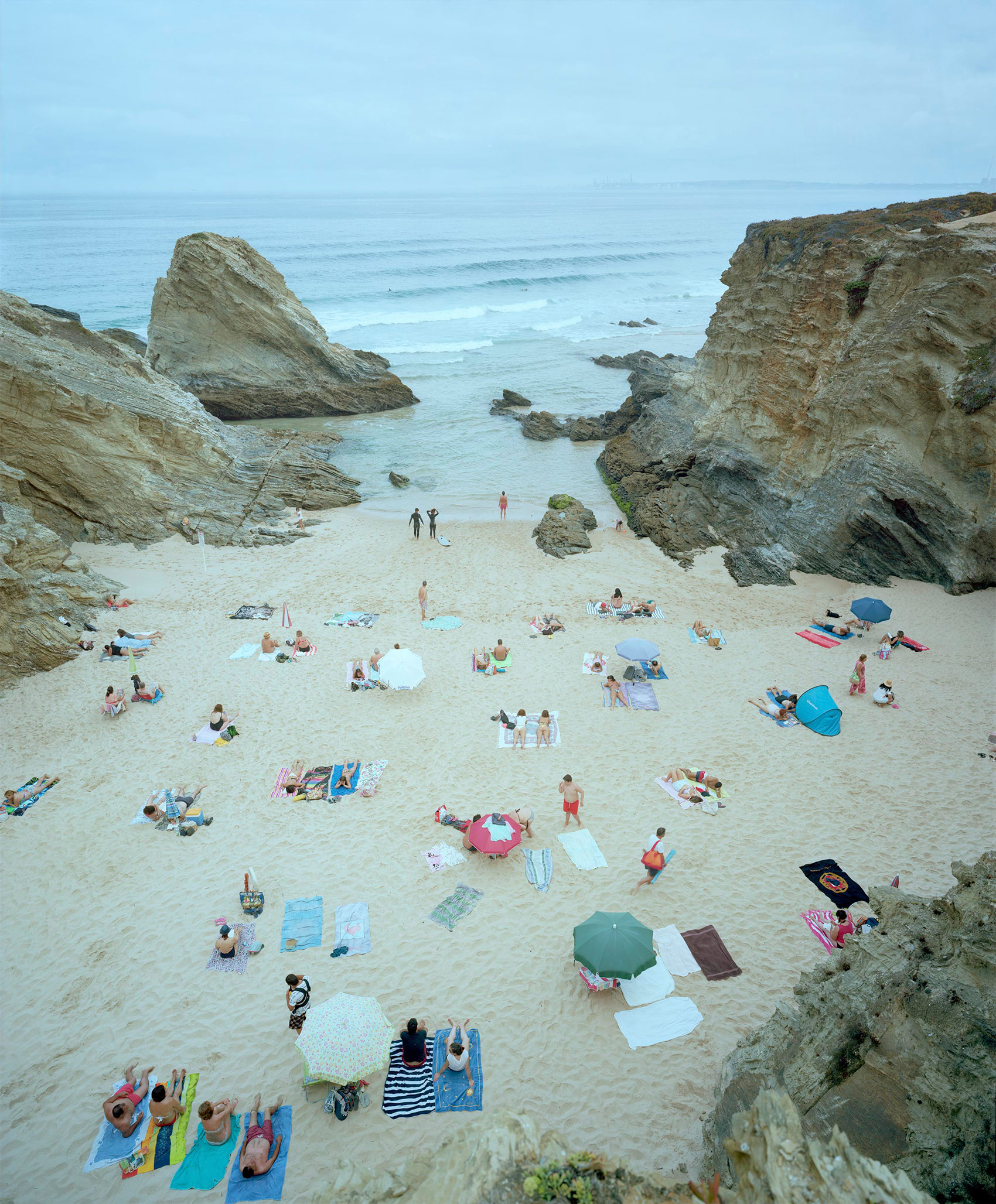 Aerial view of a crowded beach nestled between towering stratified rock formations, with numerous beachgoers scattered across pale sand among colorful umbrellas, towels, and beach equipment, calm turquoise water visible beyond.