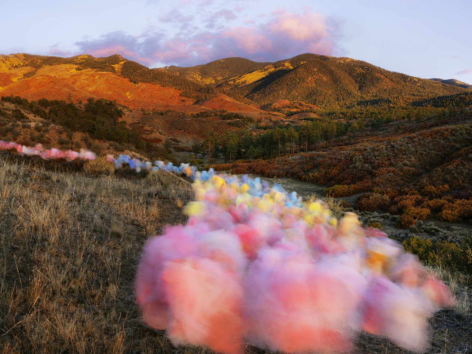 Soft, colorful tulle fabric in pink, yellow, and blue tones billows in the foreground, creating an ethereal veil across a New Mexico landscape of rust-colored hills dotted with piñon and juniper trees at golden hour.