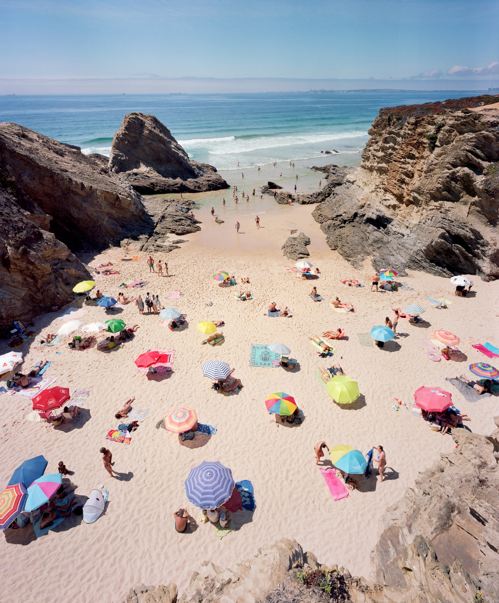 Aerial view of a crowded beach nestled between dramatic rocky outcrops, with numerous colorful umbrellas scattered across golden sand and beachgoers dispersed throughout. The composition captures the vibrant geometry of umbrellas in reds, blues, yellows, and pinks against the pale beach, with calm turquoise waters and distant shoreline visible beyond.