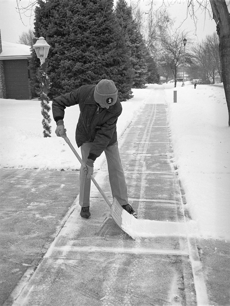 Black and white photograph of a person in winter clothing shoveling snow from a residential sidewalk, bent forward in effort. Evergreen trees, a street lamp, and snow-covered yards are visible in the background along a tree-lined street.