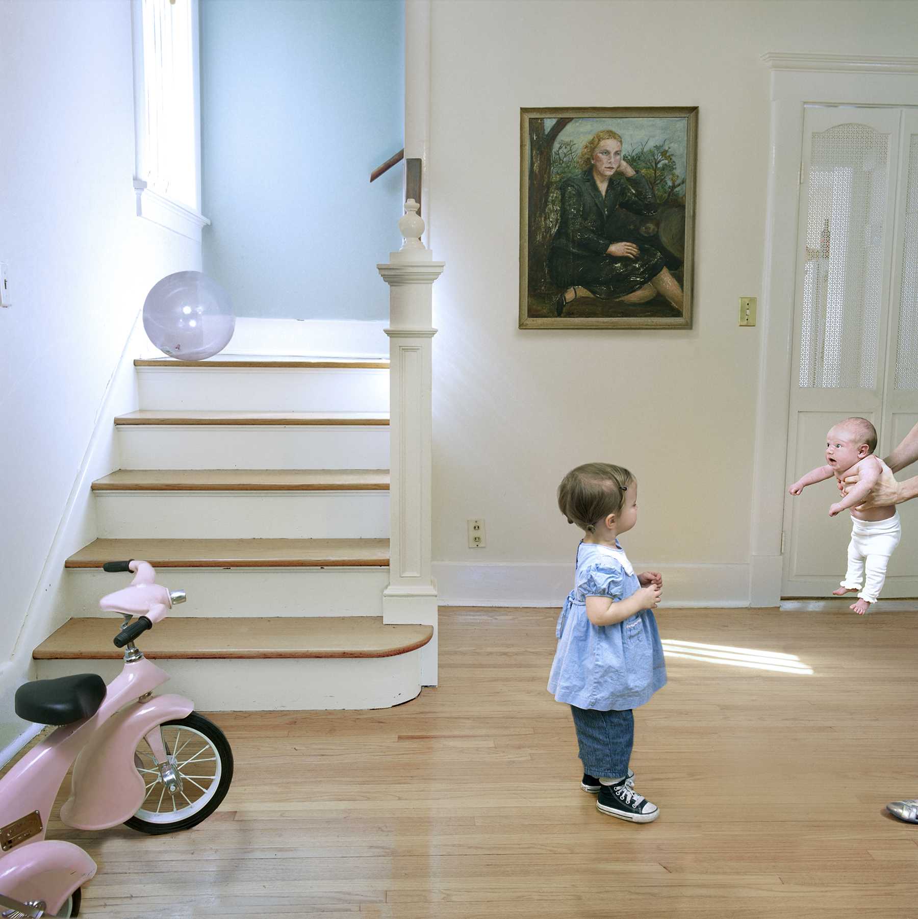 A young child in a blue shirt and jeans stands in a bright, sun-filled hallway watching a baby being held by someone on the right. A pink tricycle sits in the foreground on the left, while wooden stairs with a white banister and a framed portrait painting are visible in the background. The scene captures a moment of domestic life in a contemporary home interior.