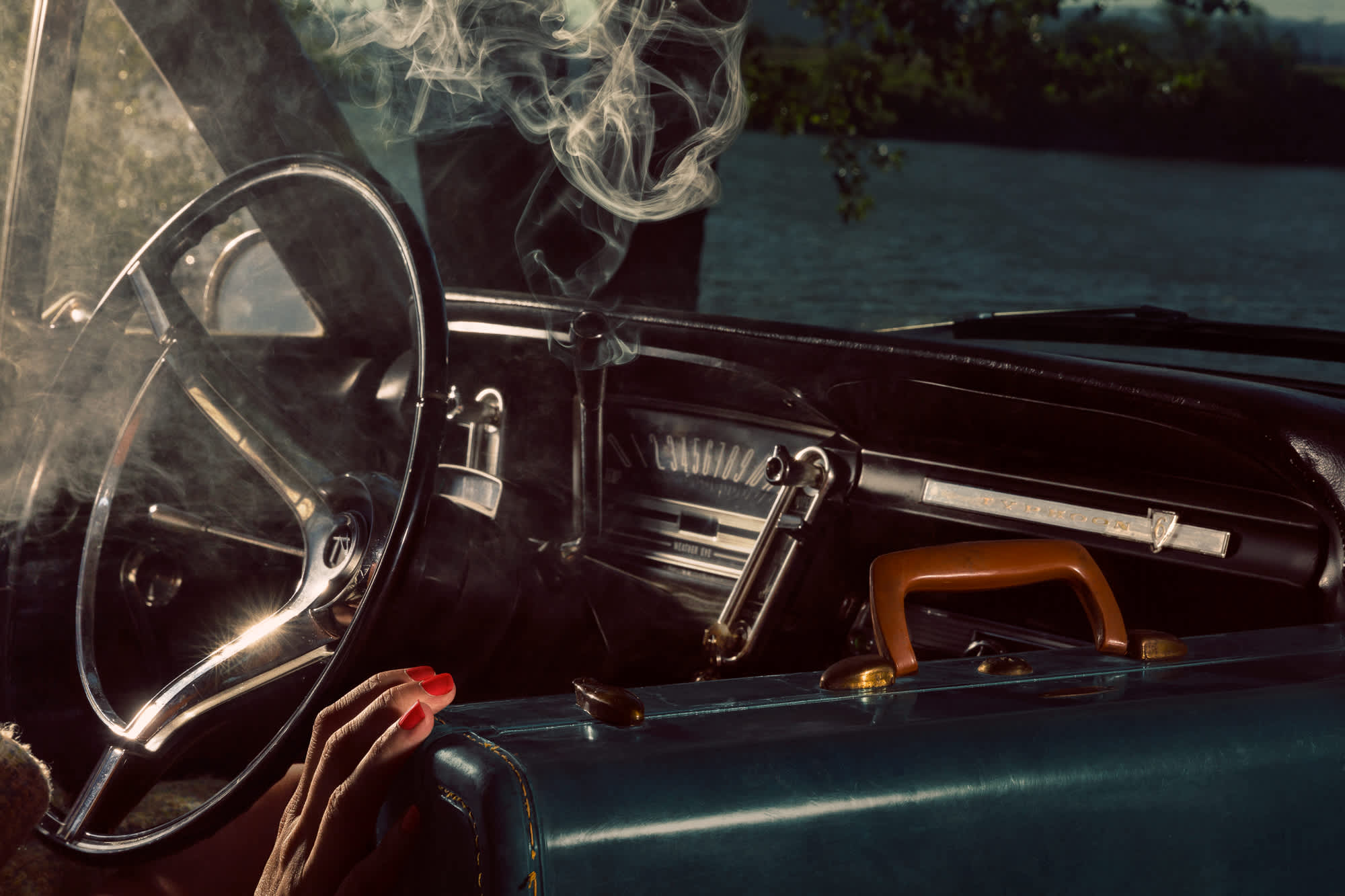 A person with red nail polish holds a cigarette inside a vintage car, smoke curling upward near the steering wheel. The dashboard and interior are rendered in deep blacks and blues, while a body of water and treeline are visible through the windshield in the background.
