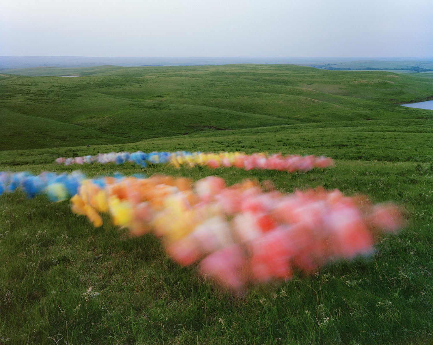 Colorful tulle fabric arranged in diagonal stripes across rolling green prairie grassland, with the layered fabric appearing in shades of blue, yellow, orange, pink, and red against the vast, undulating landscape under an overcast sky.
