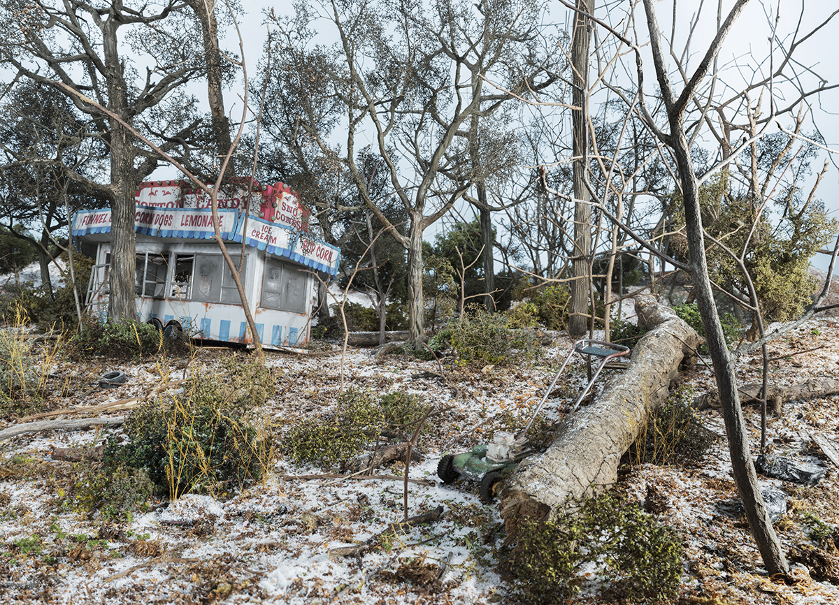 Abandoned concession stand with red and blue striping sits among bare trees and overgrown vegetation in a desolate landscape. The small structure displays faded signage for items like lemonade and ice cream, surrounded by fallen logs, scattered debris, and encroaching nature.