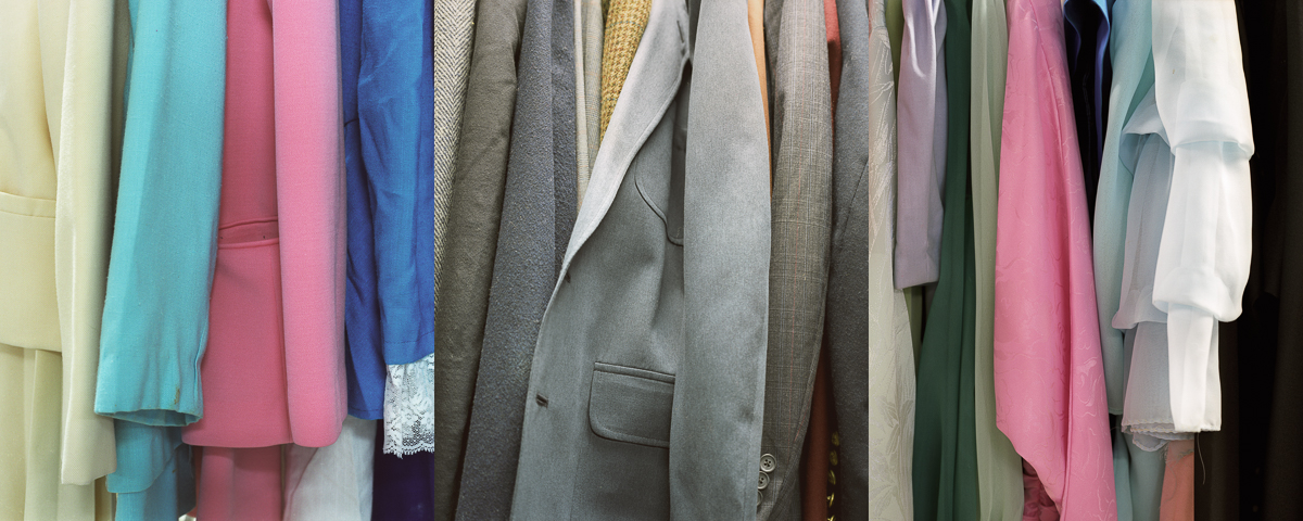 A densely packed row of hanging garments in varied colors including pastels, neutrals, and jewel tones, displayed against a neutral background. The clothing appears layered and overlapping, showcasing different fabrics and textures in shades of pink, blue, gray, green, and white.