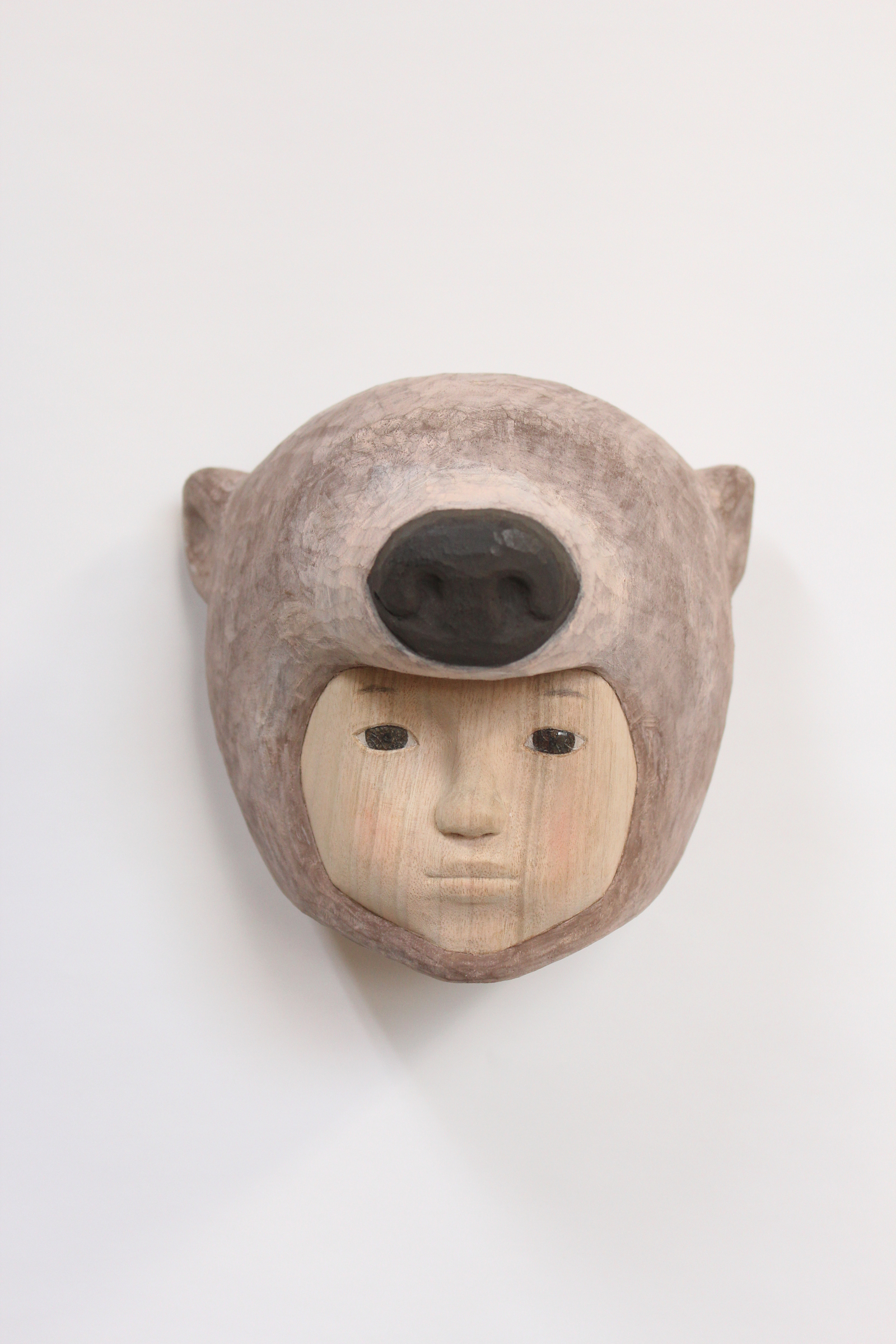 Carved wooden mask depicting a human face with closed eyes and neutral expression, framed by a rounded animal head with pointed ears and a large dark oval opening where the eyes would be. The lighter wood face contrasts with the darker weathered tones of the surrounding animal form.