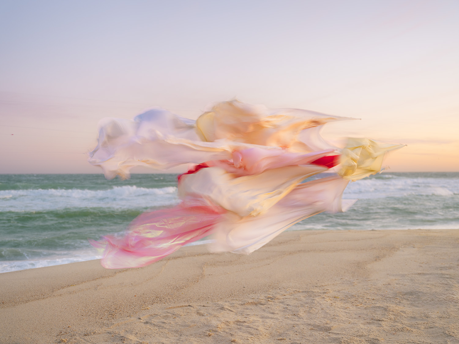 Flowing silk fabrics in cream, pink, red, and yellow billow across a sandy beach at sunset, with soft waves and ocean visible in the soft-focus background.