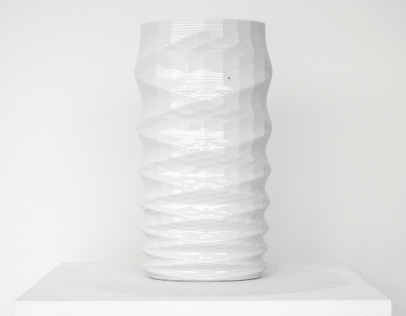 A tall cylindrical porcelain vessel with a twisted, ribbed surface creating a helical pattern. The piece is rendered entirely in white glaze with subtle shadows emphasizing its spiraling form, displayed against a neutral background on a white platform.