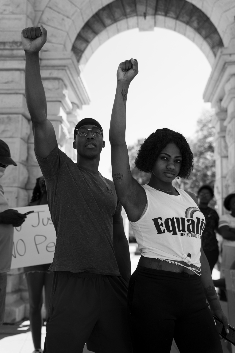 Black and white photograph of two people with raised fists standing beneath a classical archway, with protest signs visible in the background. The woman on the right wears a shirt reading "Equality," while architectural elements and other demonstrators frame the composition.