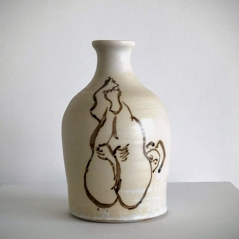 A cream-colored ceramic bottle with a tapered neck features a gestural brown line drawing of a stylized nude figure on its rounded body. The minimalist figural sketch, rendered in an expressive manner, contrasts with the vessel's smooth, functional form.