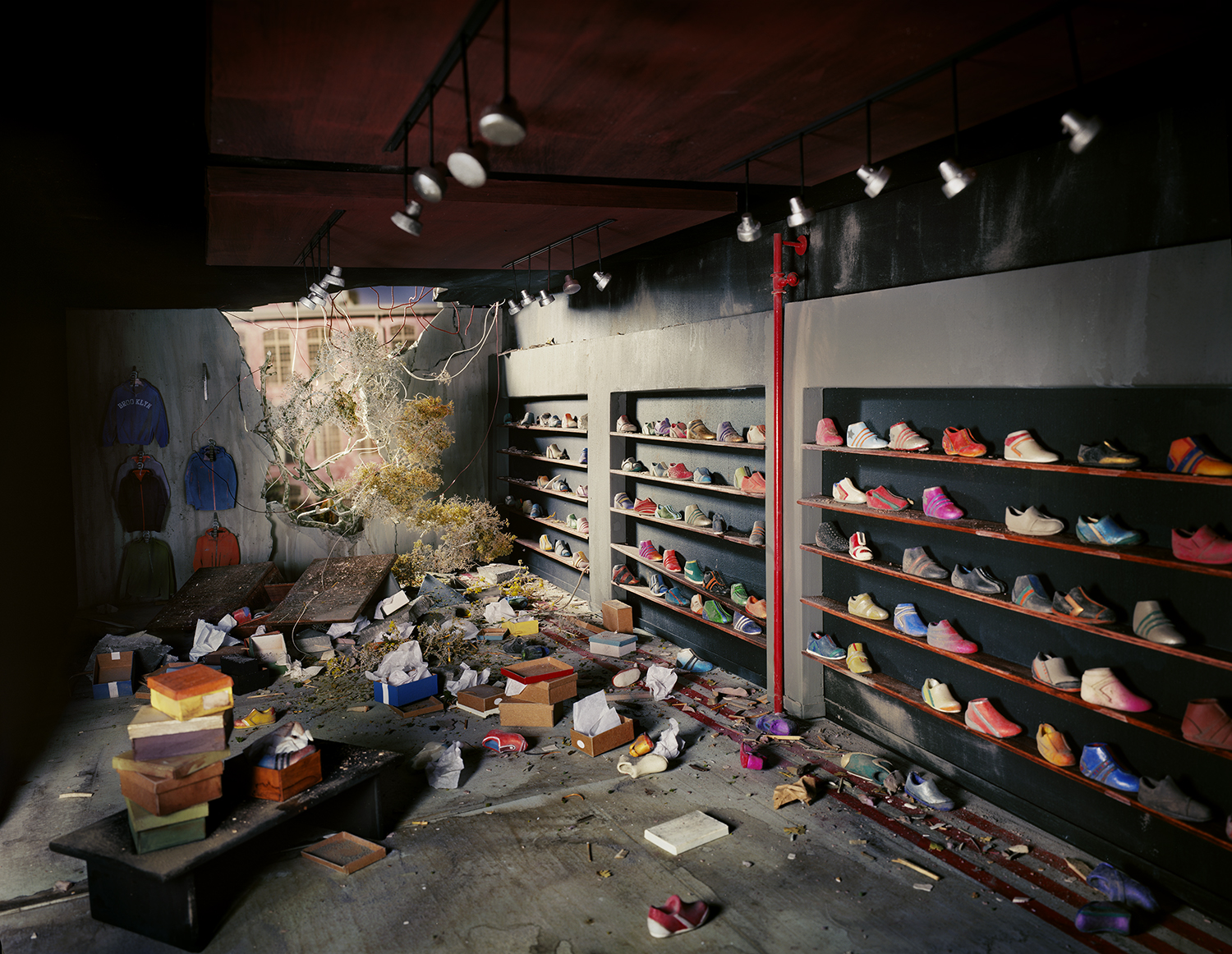 An indoor retail space lies in ruins, with colorful sneakers displayed on black wall-mounted shelves while debris, overturned boxes, and scattered merchandise cover the concrete floor. Bare branches protrude from the wreckage beneath overhead track lighting, creating a surreal juxtaposition of commercial order and catastrophic destruction.