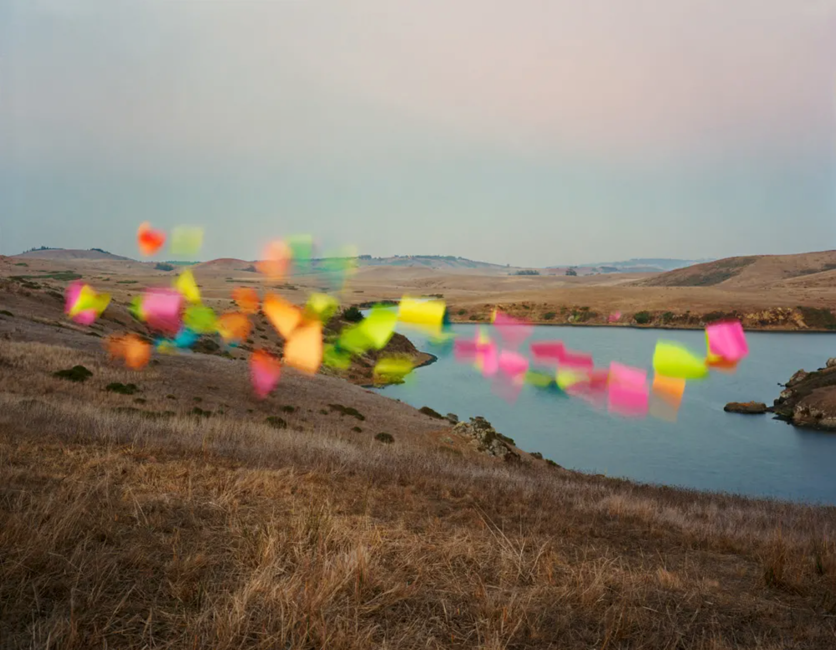 Arid hillside landscape with dry golden grasses overlooks a calm blue reservoir, with rolling brown hills visible in the distance. Vibrant, translucent colored shapes—hot pink, yellow, orange, green, and cyan—float across the scene, obscuring portions of the natural vista.
