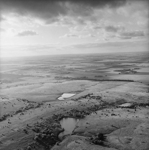 Aerial black and white photograph of a vast, flat landscape dotted with scattered trees and several bodies of water reflecting light against darker fields. Dark storm clouds gather across the expansive sky above the horizon.
