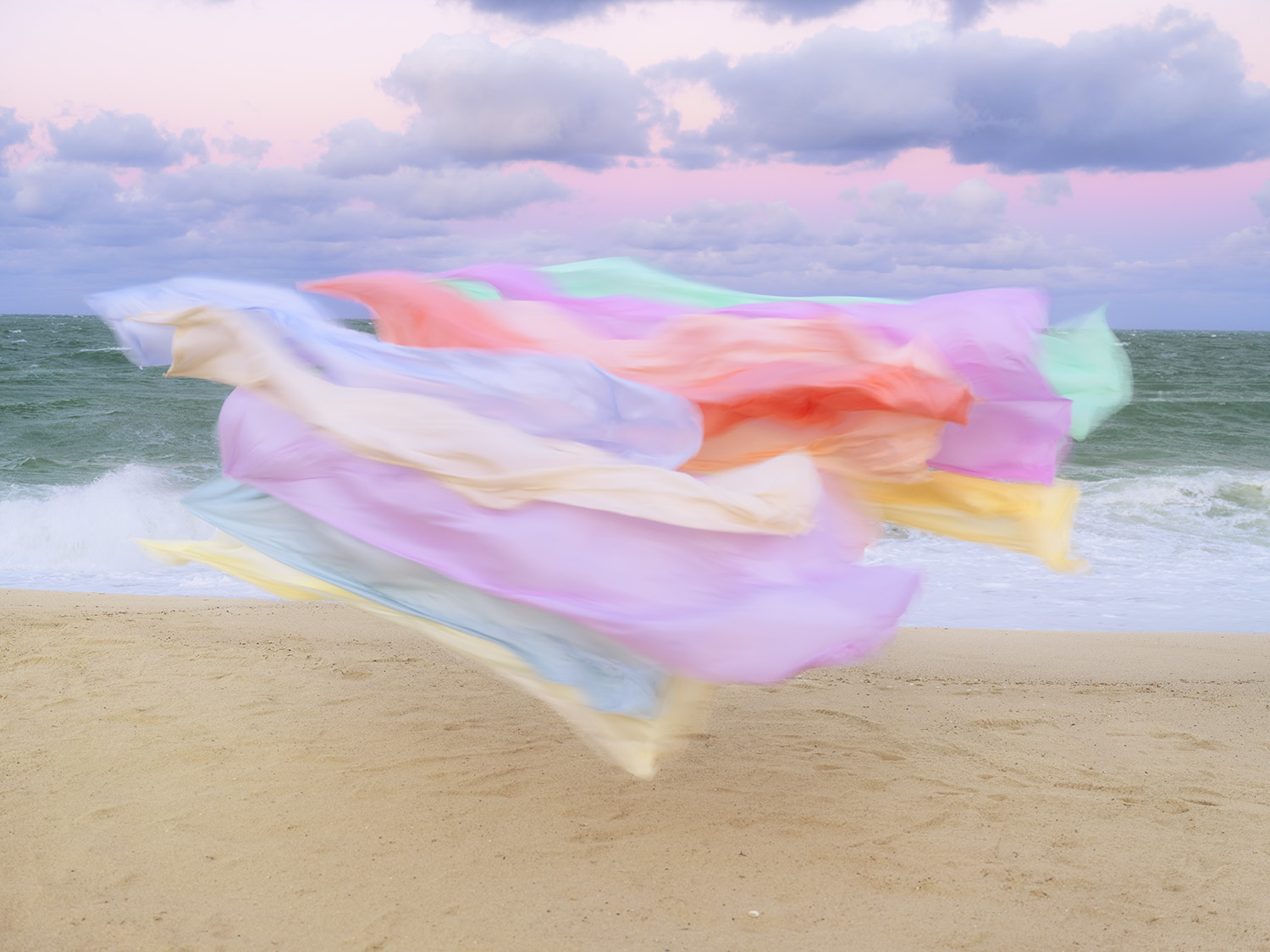 Colorful silk fabric in pastel hues of pink, lavender, yellow, coral, and mint green billows dramatically across a sandy beach, with ocean waves and cloudy sky visible in the background.