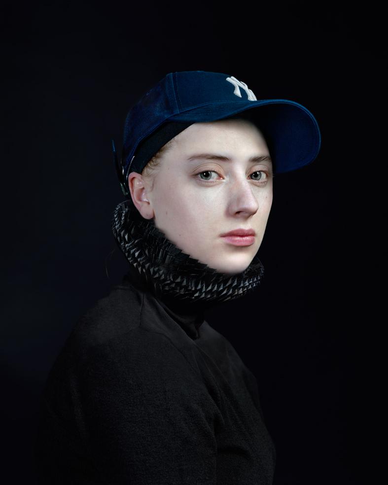 Portrait of a young person facing slightly to the right against a black background, wearing a dark knitted turtleneck and navy blue baseball cap with a white logo. The subject has pale complexion, defined features, and dark hair pulled back, rendered in sharp photographic detail with dramatic lighting that emphasizes facial contours.